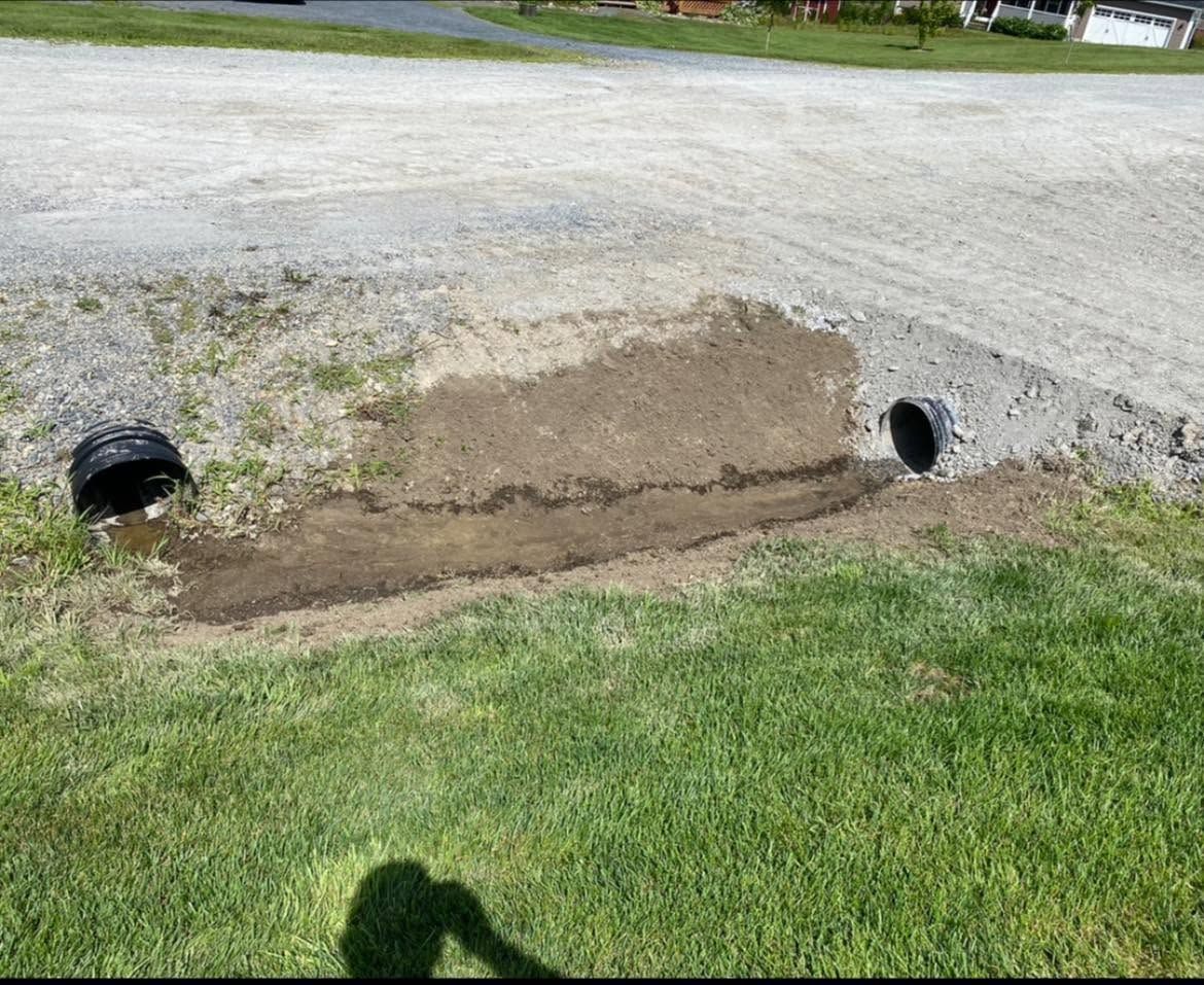 Eroded roadside ditch with exposed drainage pipes and muddy soil beside a gravel road.