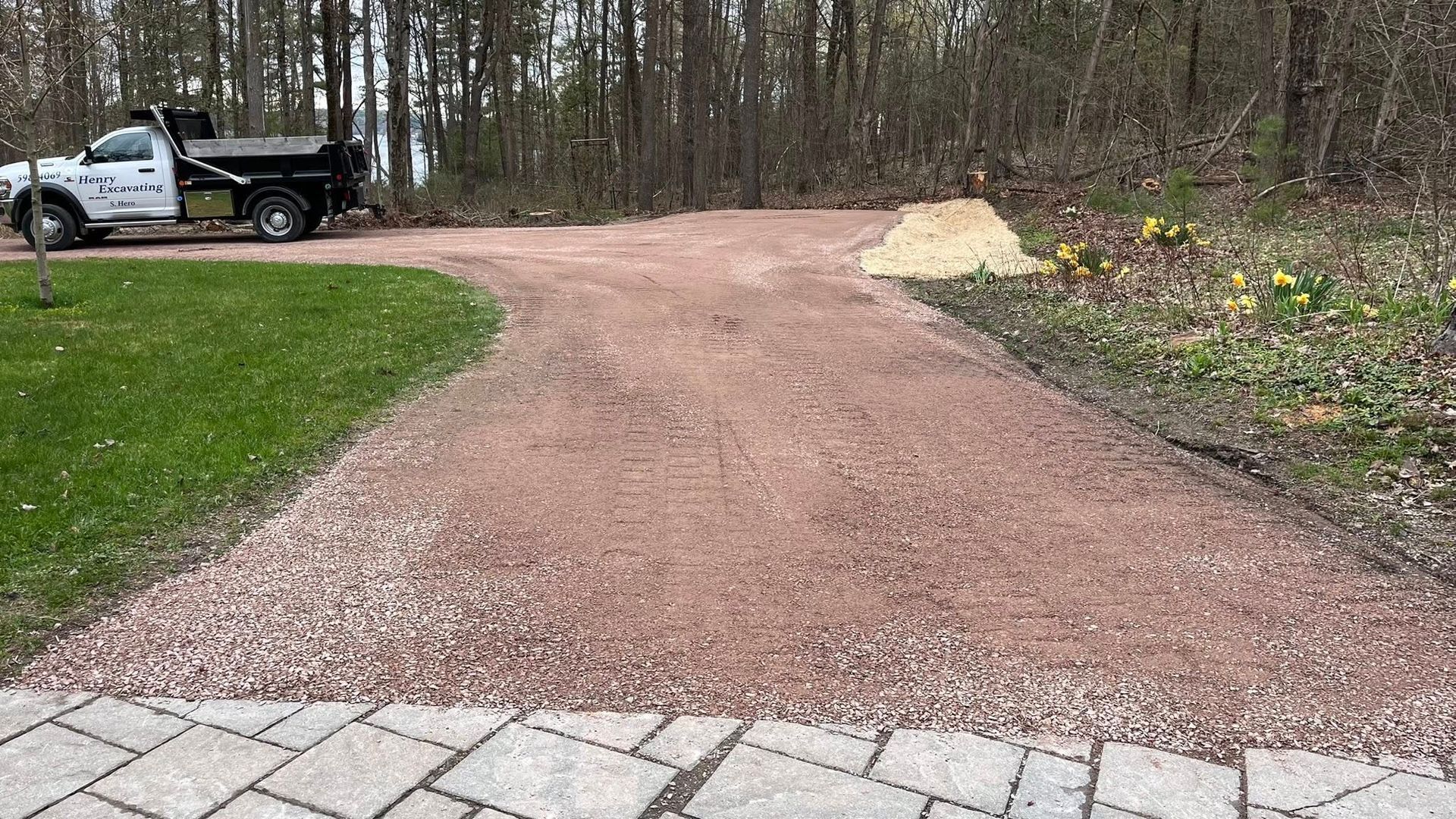 Gravel driveway under construction beside grass, with a pickup truck and pile of gravel near a wooded area.
