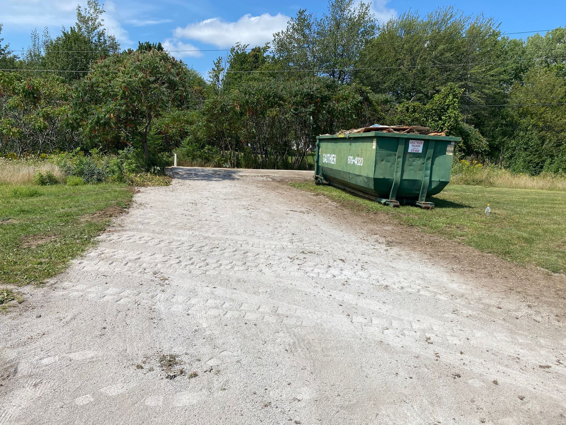 Gravel driveway beside a green dumpster, with grass and trees under a blue sky