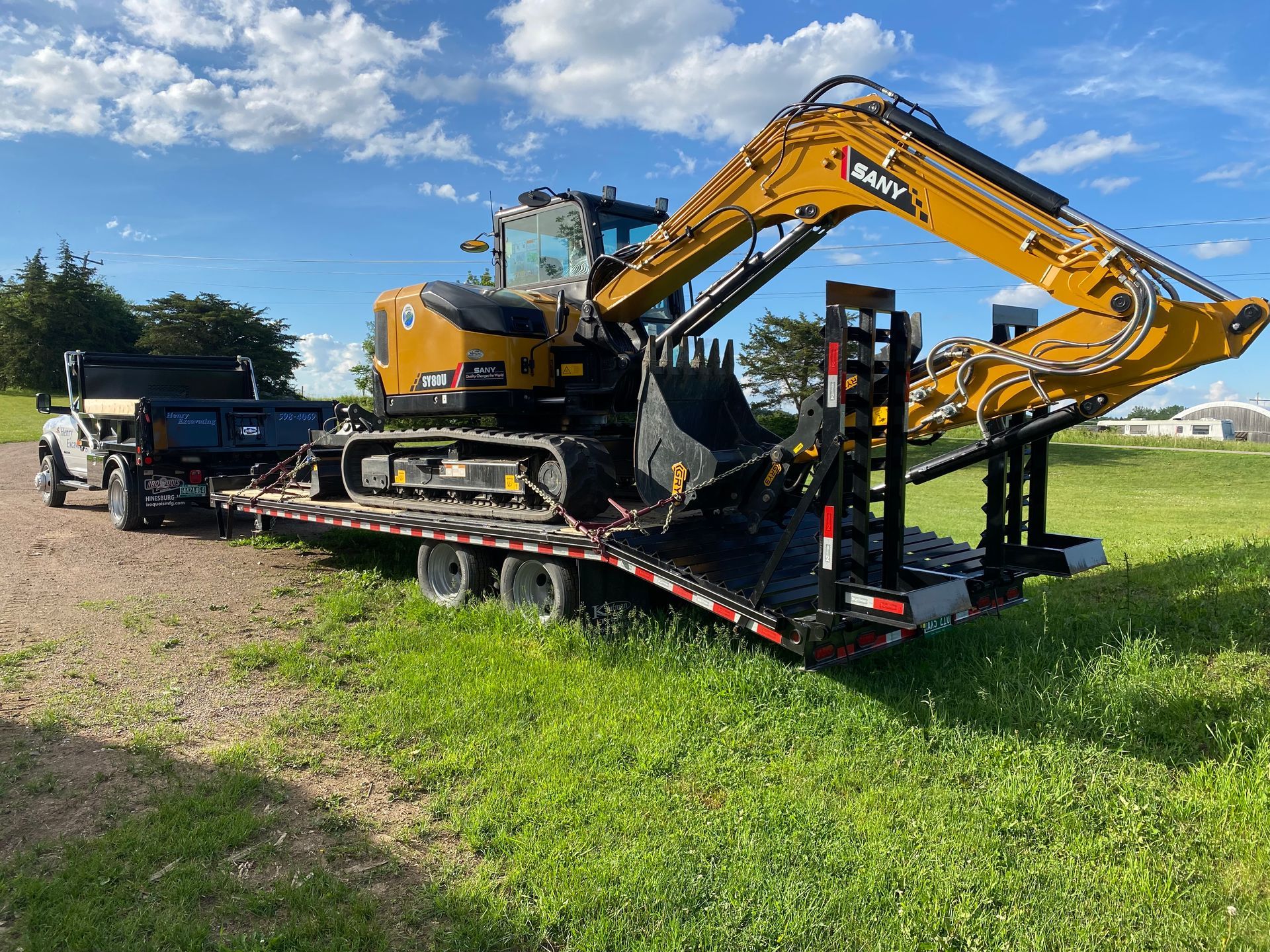 Yellow excavator loaded on a flatbed trailer in a grassy lot under a blue sky
