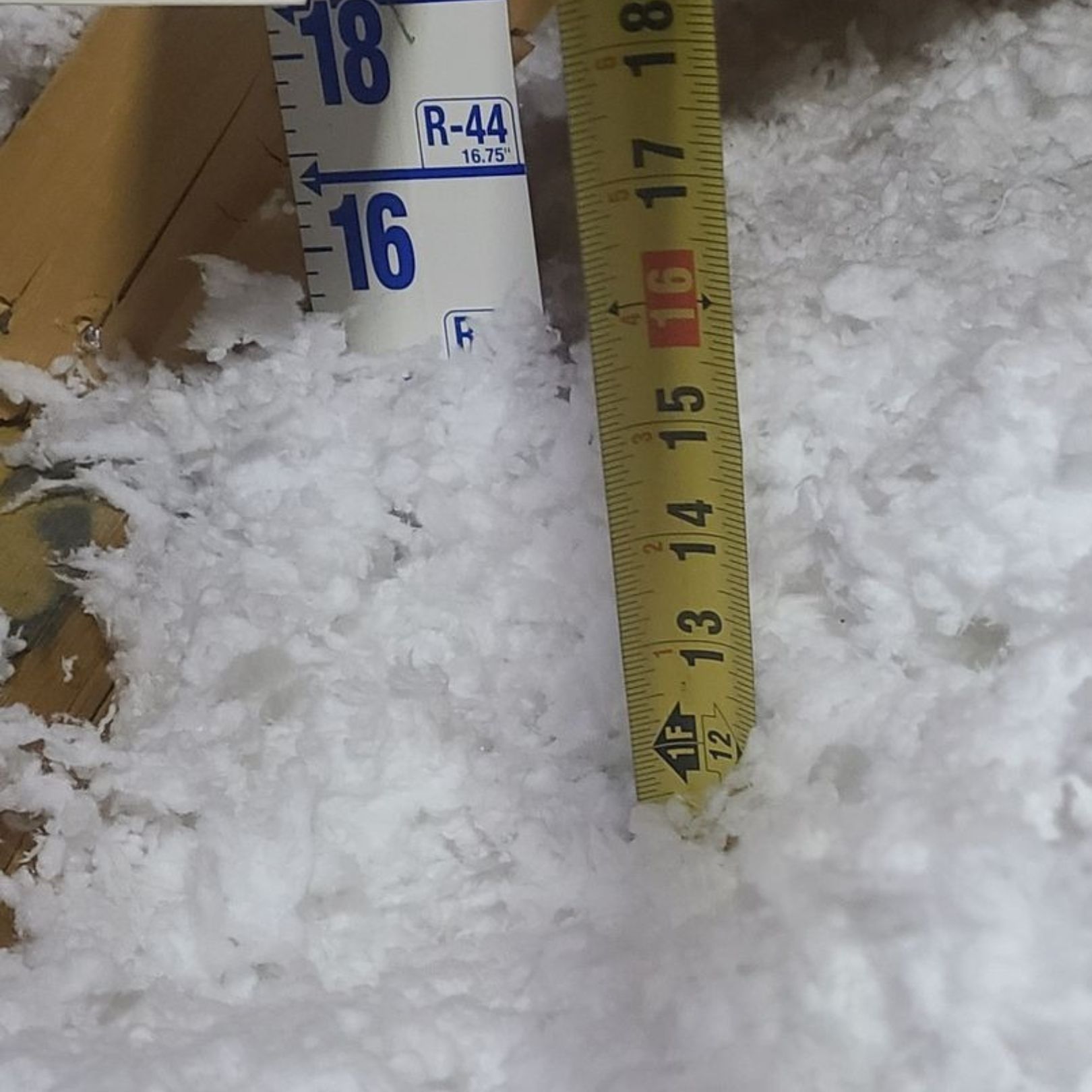 A tape measure indicates the depth of white blown-in attic insulation is approximately 12.5 inches deep.