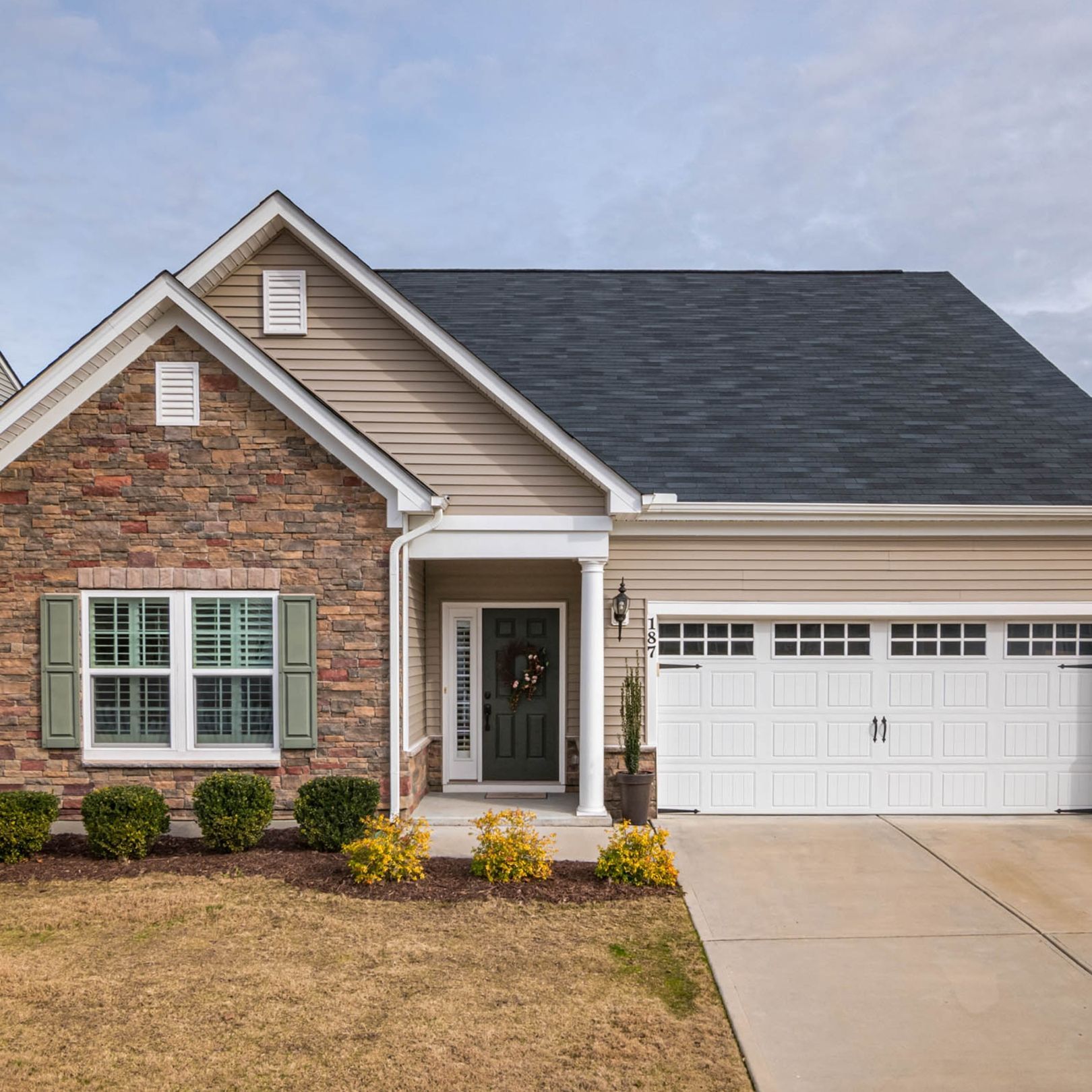 A tan, single-story house with a stone facade, dark roof, white garage door, and a small front yard with shrubs.