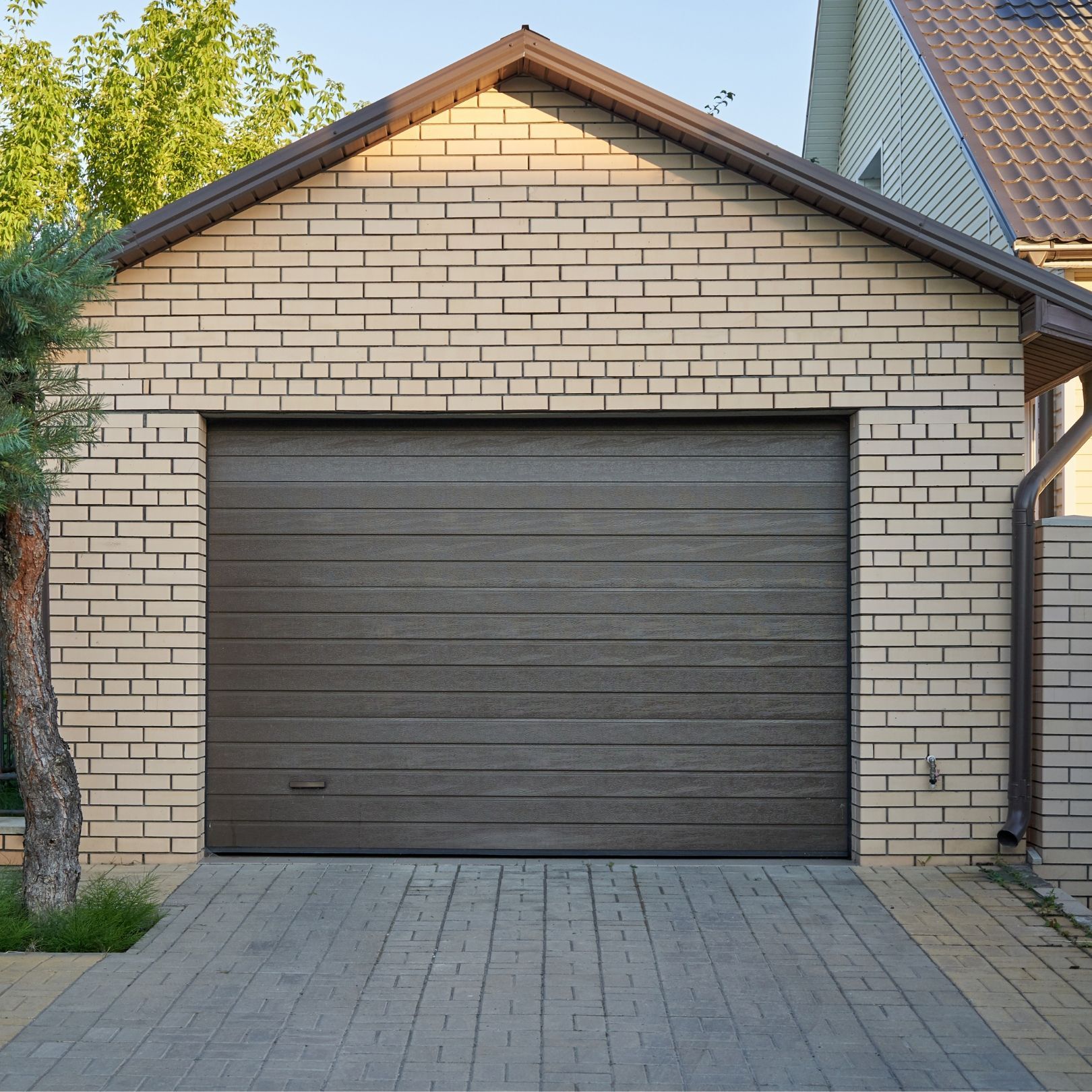 A brick garage with a brown sectional door, set against a paved driveway and a clear blue sky.