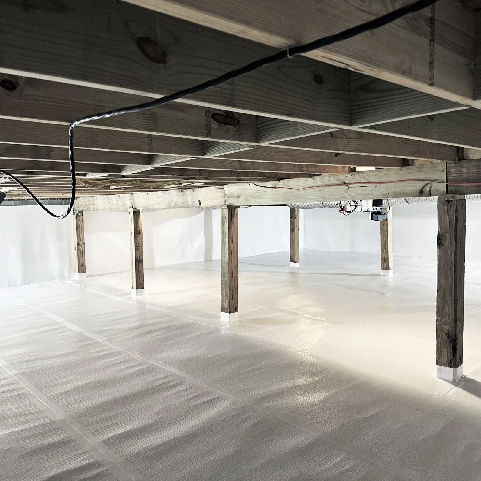 A crawl space with wooden support beams and floor joists above a clean, white, vapor-barrier-lined floor.
