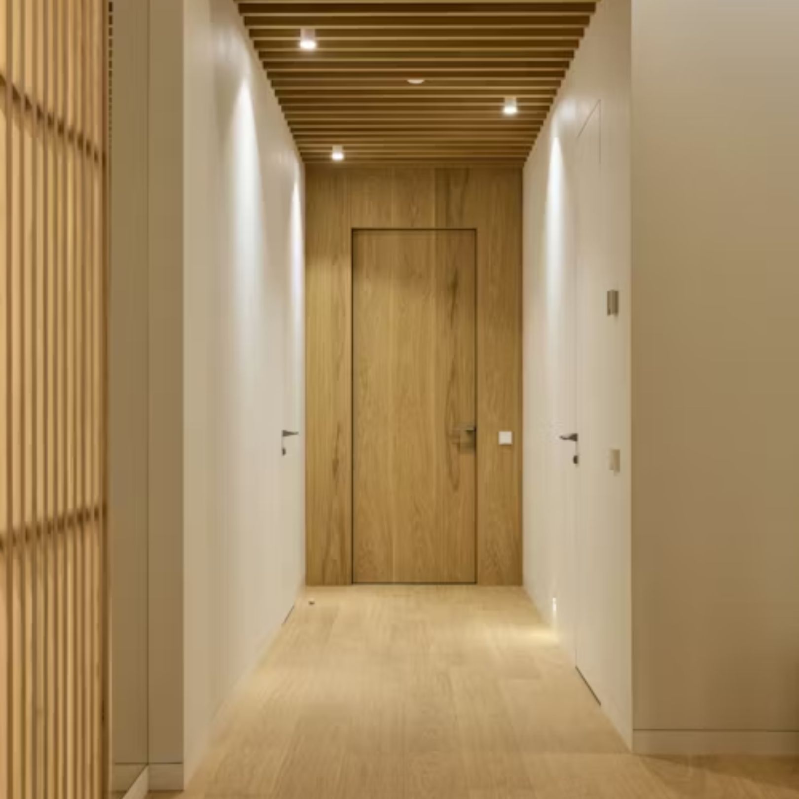 A hallway featuring light wood flooring, a wooden slatted ceiling, a central wooden door, and minimalist white walls.