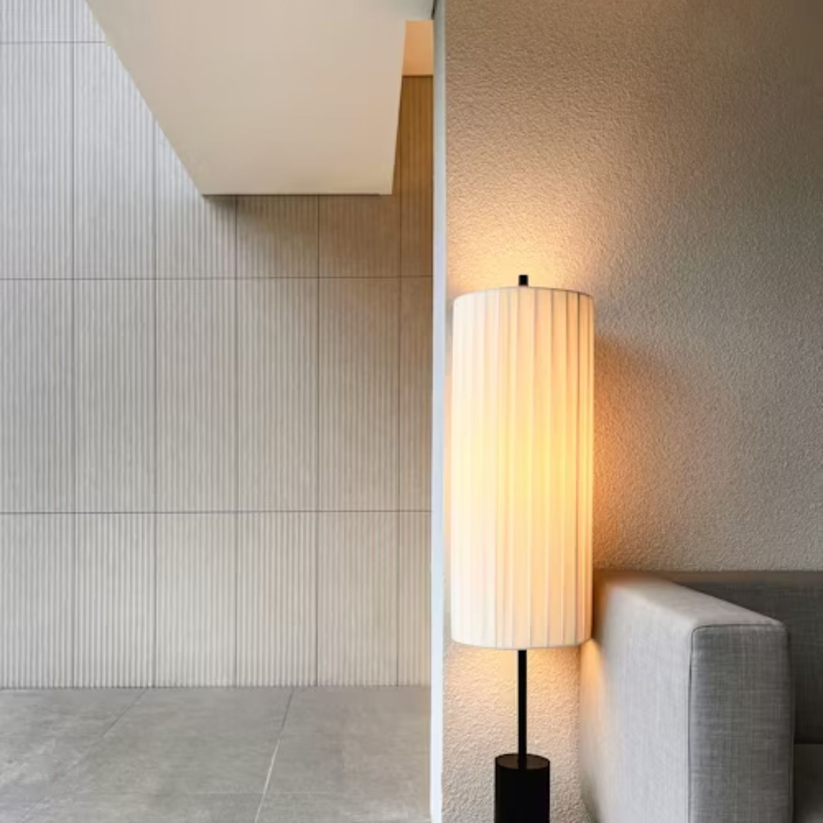 A floor lamp with a textured, pleated cylindrical shade illuminates a corner near a gray sofa and a slatted wall.