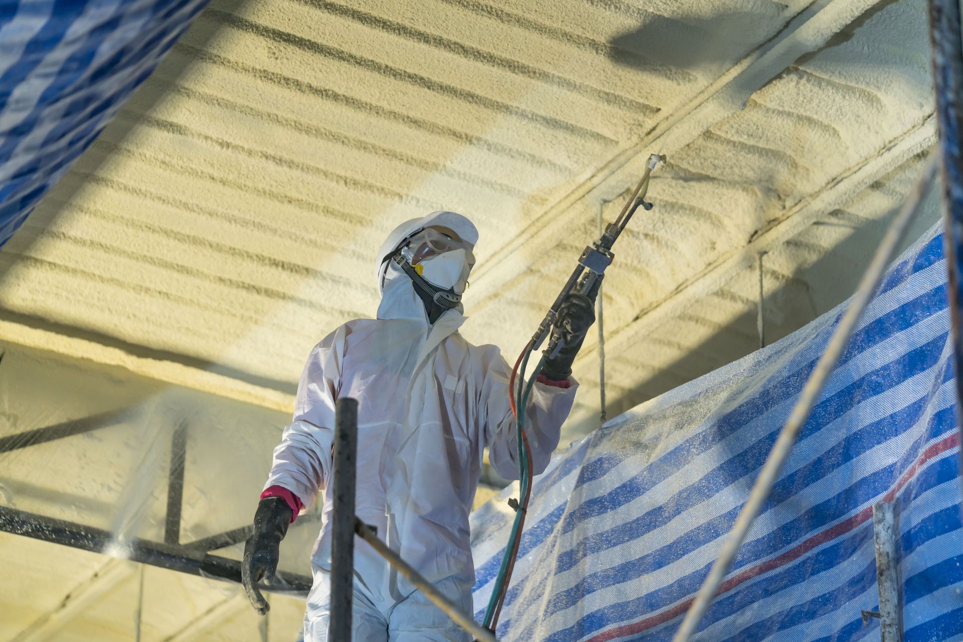 A worker in a white protective suit and mask sprays insulation foam onto the ceiling of a building.