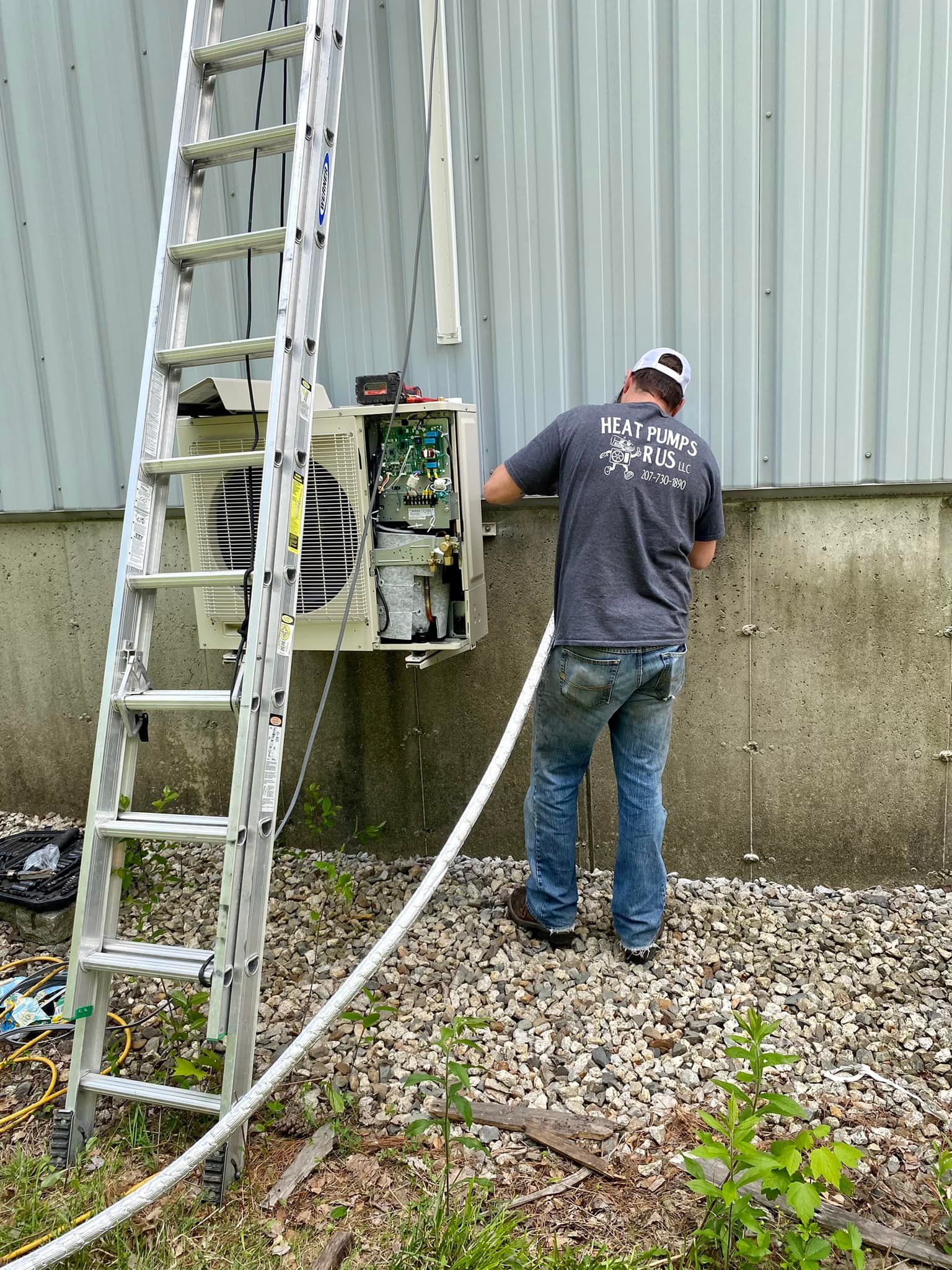 A person in a gray shirt stands on gravel, servicing an outdoor air conditioning unit mounted on a gray metal wall.