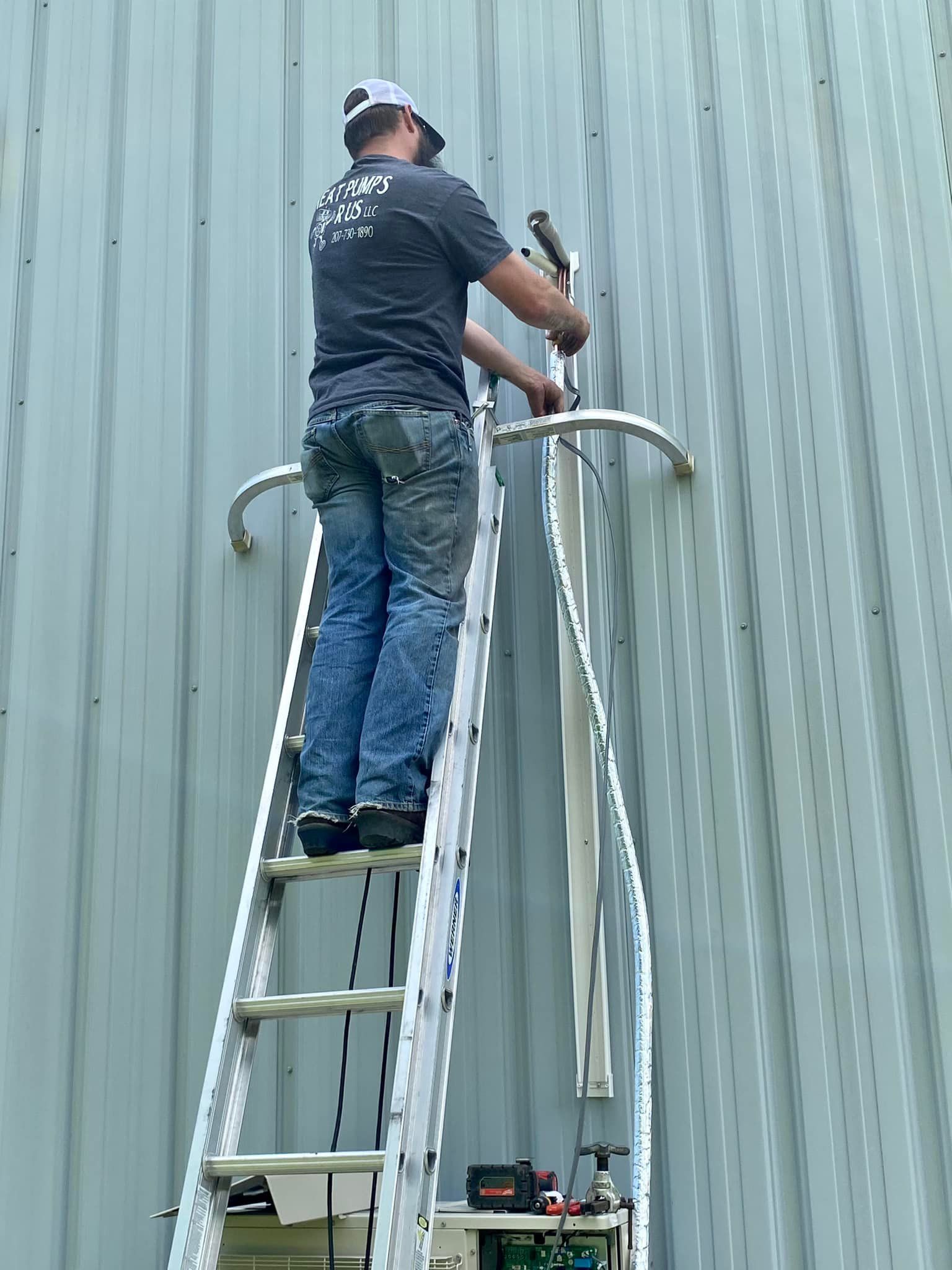 A technician on a tall ladder works on equipment mounted to a light-colored, vertically ribbed metal wall.