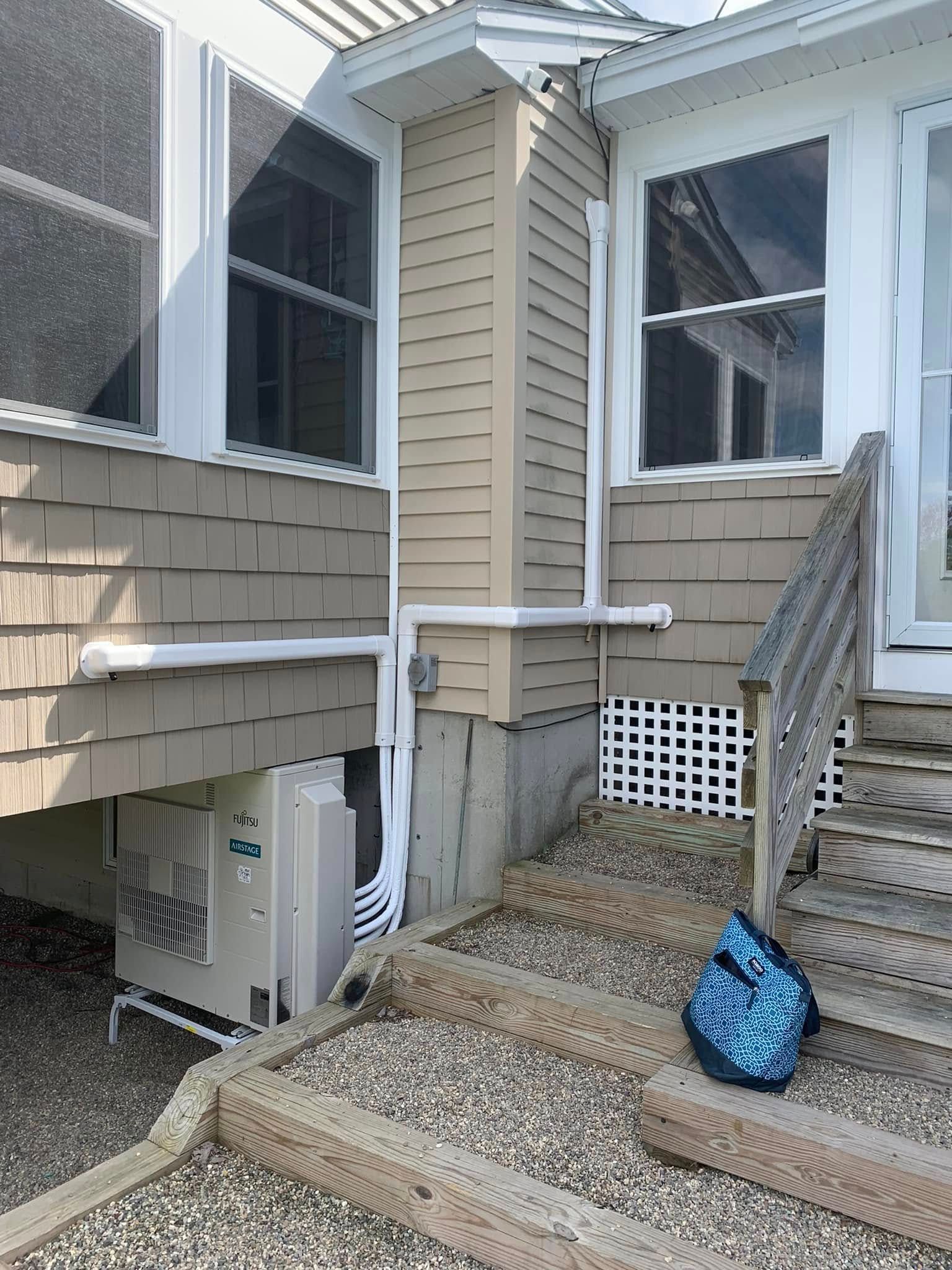 Exterior of a house with tan siding, stairs leading to a doorway, and a white HVAC unit connected by external pipe covers.
