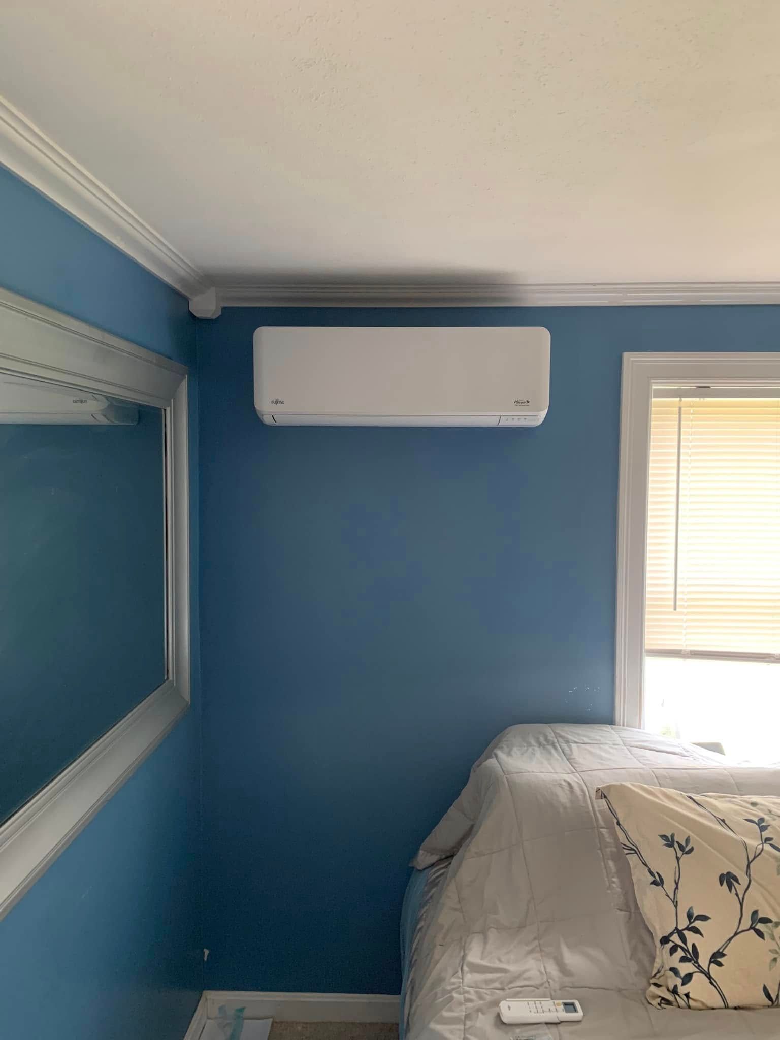 A white ductless mini-split air conditioner mounted on a blue wall between a mirror and a window with blinds.