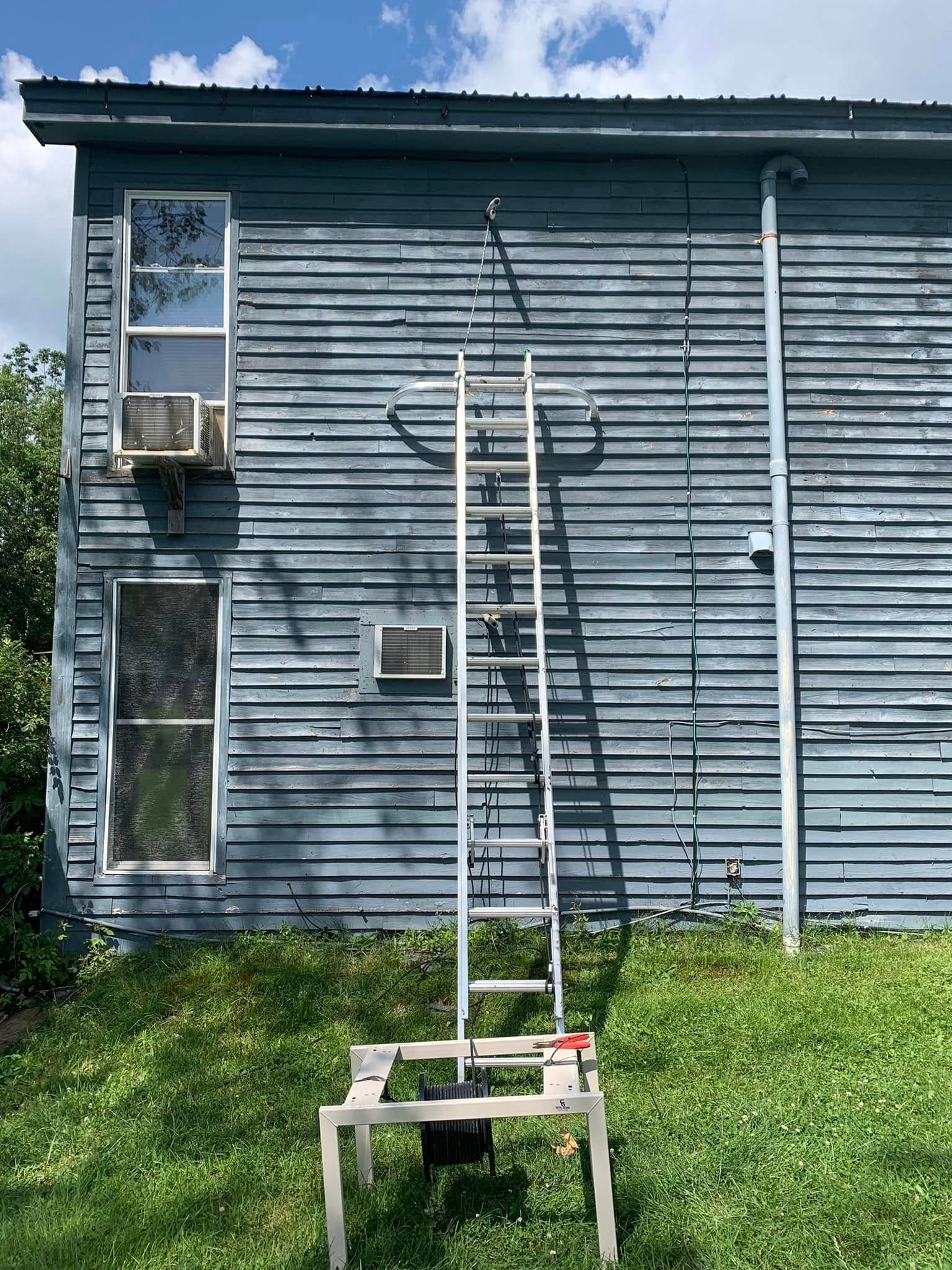An extension ladder stands against the weathered blue, wood-sided wall of a two-story building on a grassy lawn.