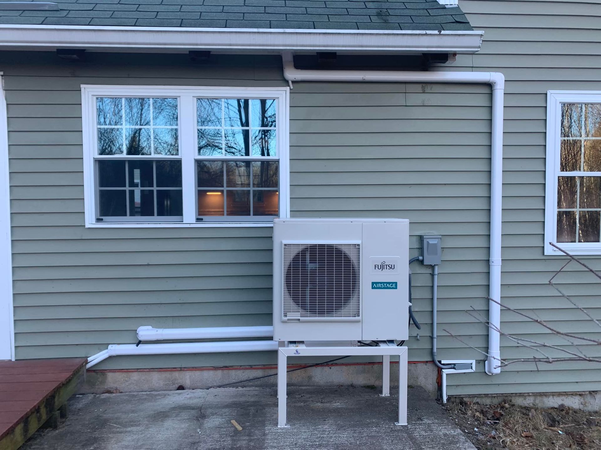 An outdoor mini-split heat pump unit installed on a white metal stand against the light green siding of a house.