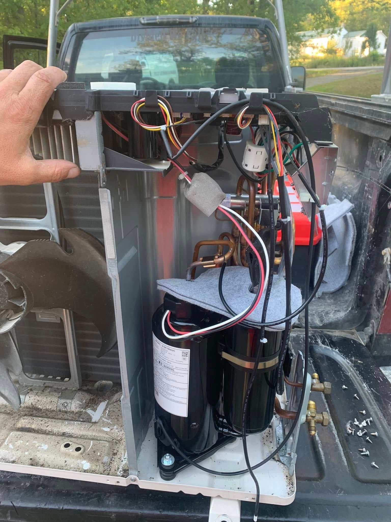 A hand holds the frame of an open outdoor HVAC condenser unit, revealing the compressor and internal wiring in a truck bed.