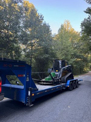 Skid steer loader on a blue trailer in a wooded area.