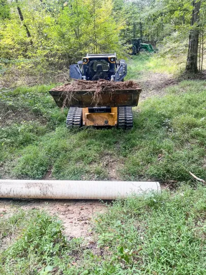 A small bulldozer with a dirt-filled bucket above a corrugated culvert pipe in a grassy wooded area.
