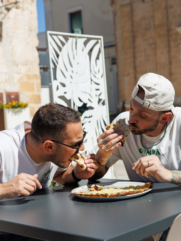 Due uomini mangiano la pizza seduti a un tavolo all'aperto. Indossano entrambi camicie bianche e occhiali da sole. Luce solare intensa.