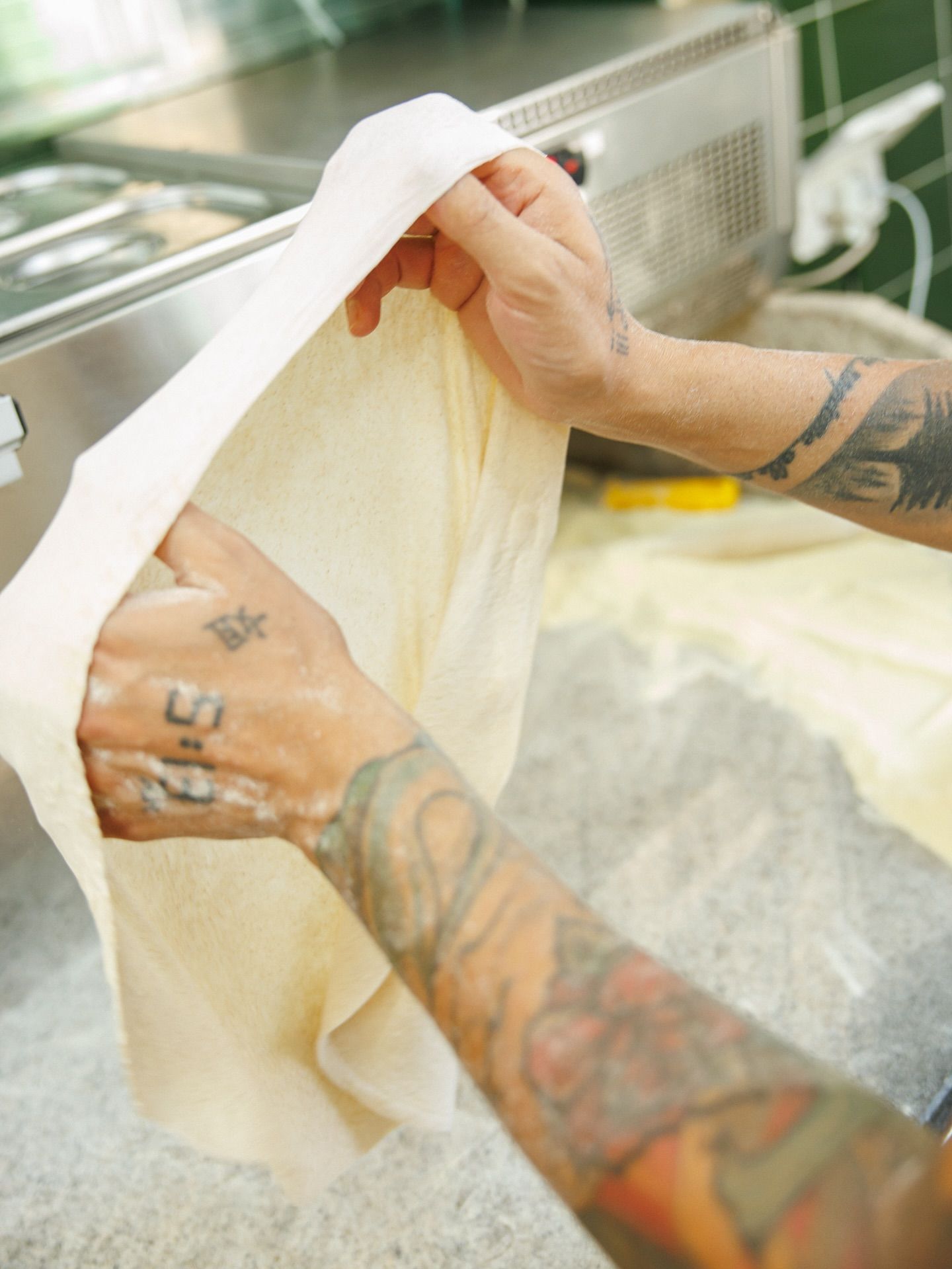 Mani che stendono l'impasto in cucina. Tatuaggi sulle braccia.