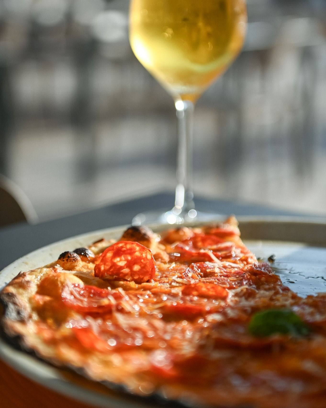 Una fetta di pizza con salame piccante e un bicchiere di vino in un ambiente soleggiato all'aperto.