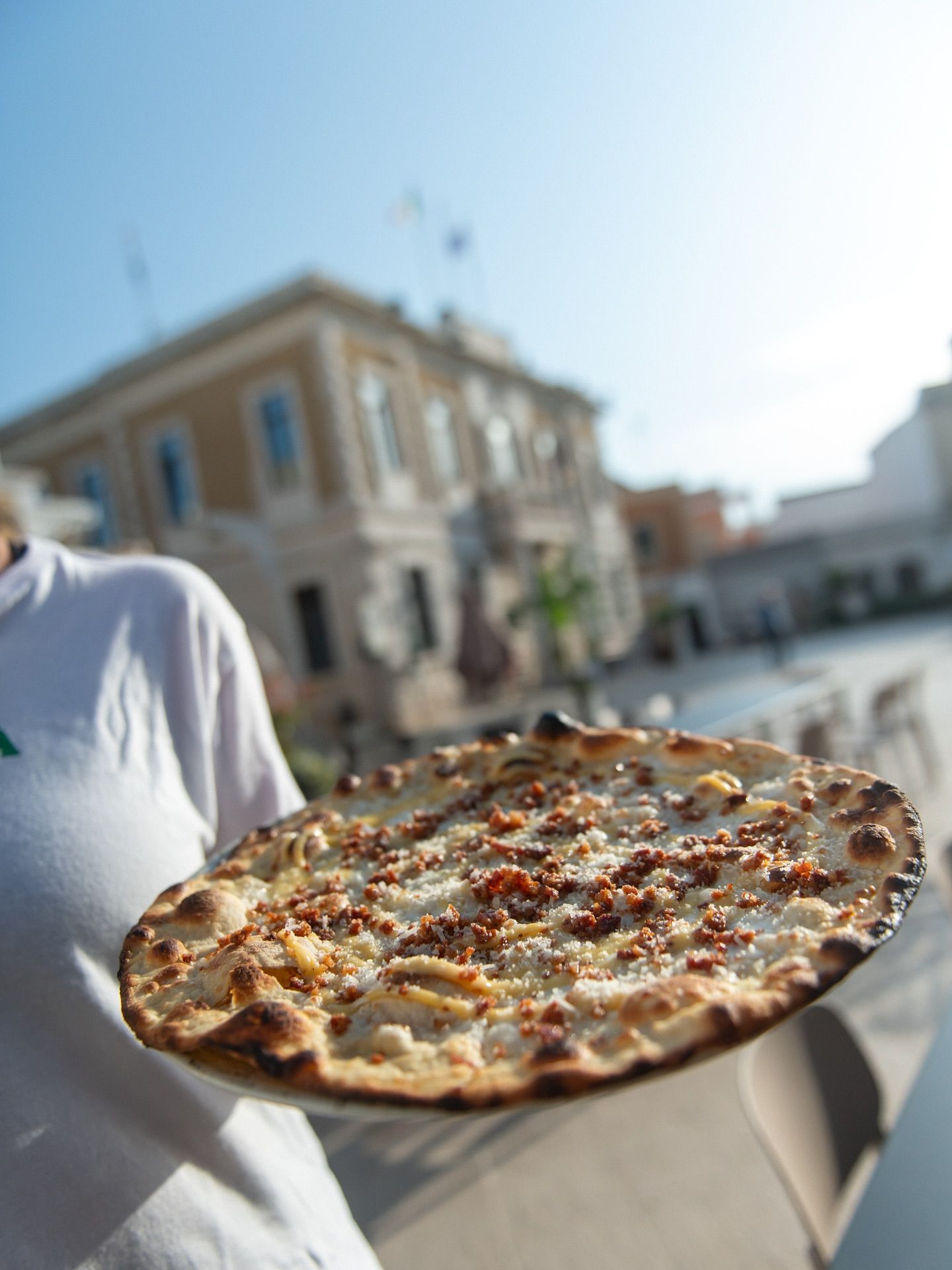 Persona che tiene in mano una pizza rotonda all'aperto, di fronte a un edificio con una piazza.