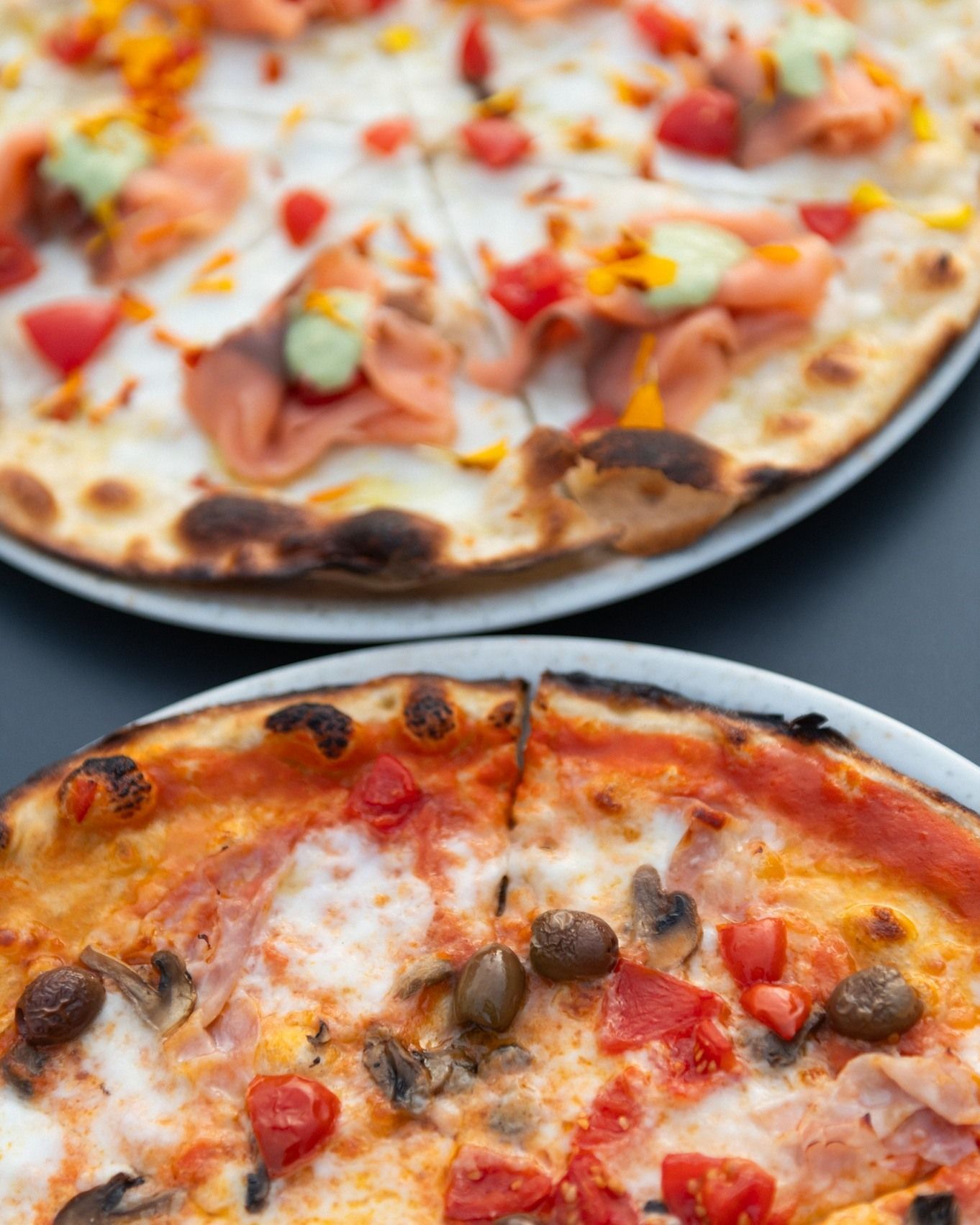 Due pizze: una al salmone, l'altra con pomodori, olive e condimenti, servite su piatti bianchi.