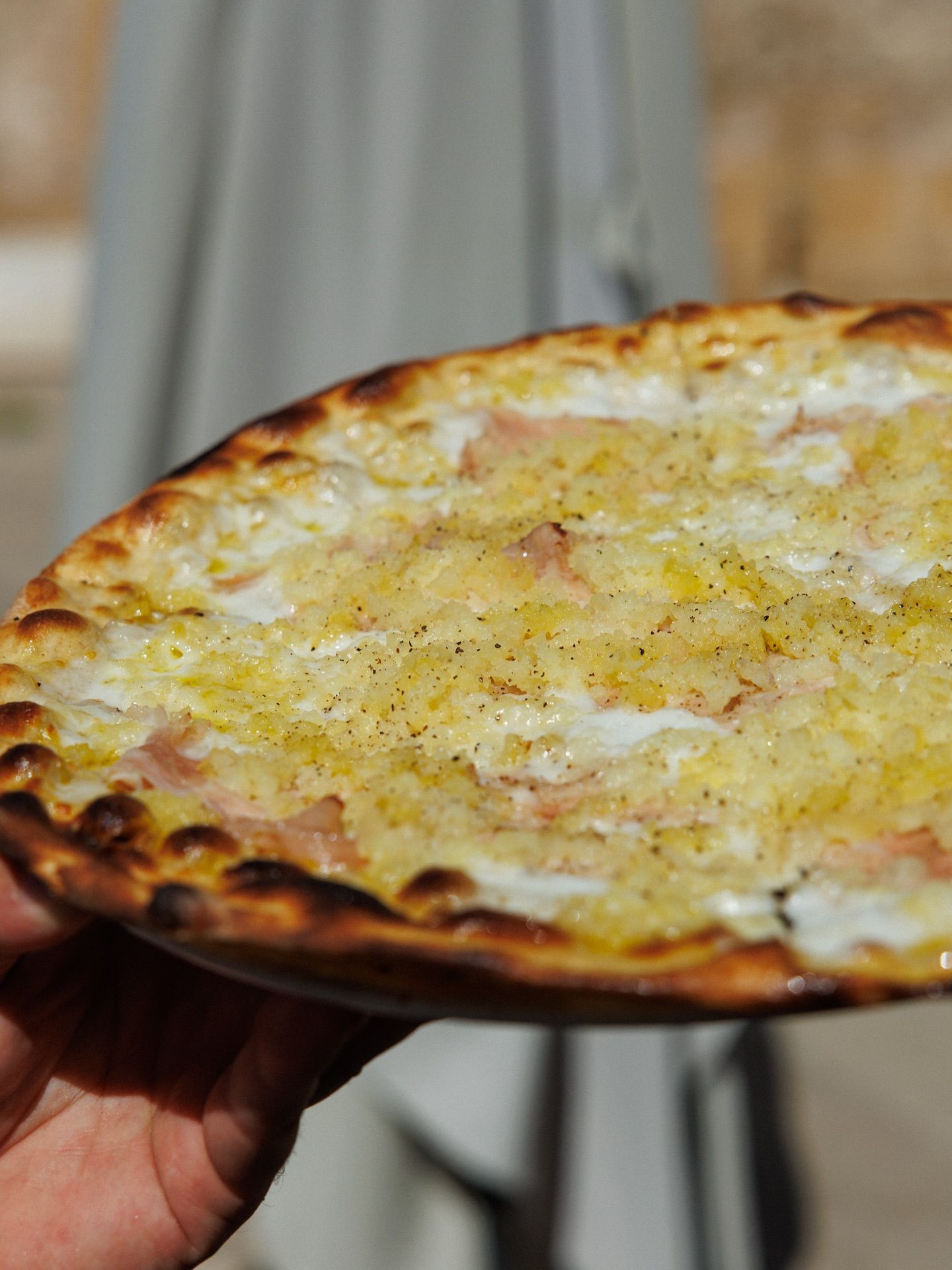 Mano che tiene una pizza rotonda con un condimento chiaro e una crosta scura, leggermente bruciacchiata.