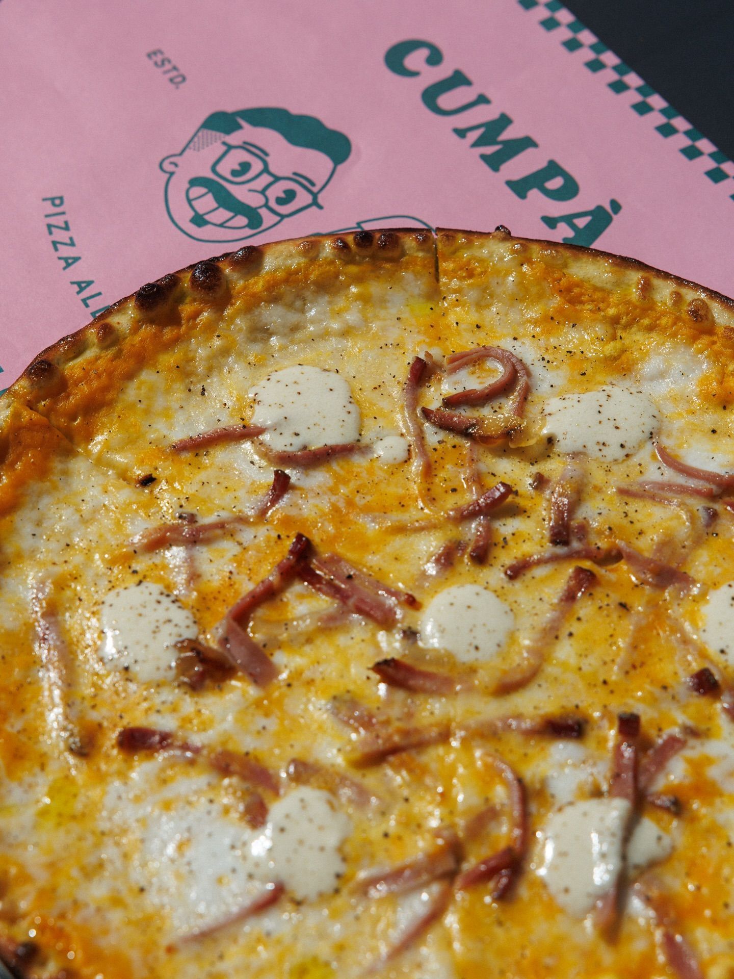 Primo piano di una pizza con prosciutto e formaggio cremoso su una scatola rosa con un logo a fumetti.