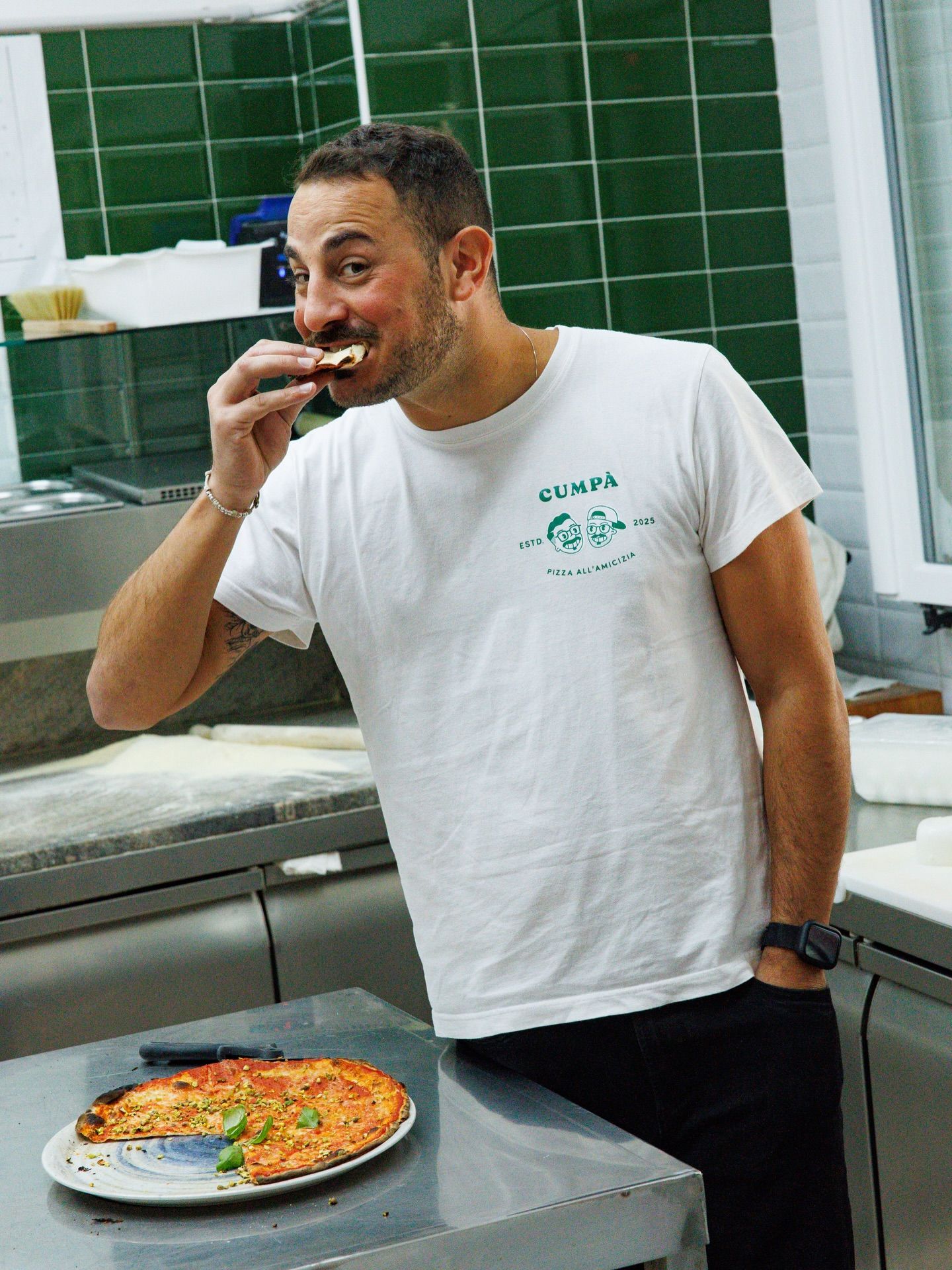 Uomo che mangia una fetta di pizza in cucina. Pizza sul bancone; camicia bianca, pantaloni neri, guarda la fetta con un sorriso.