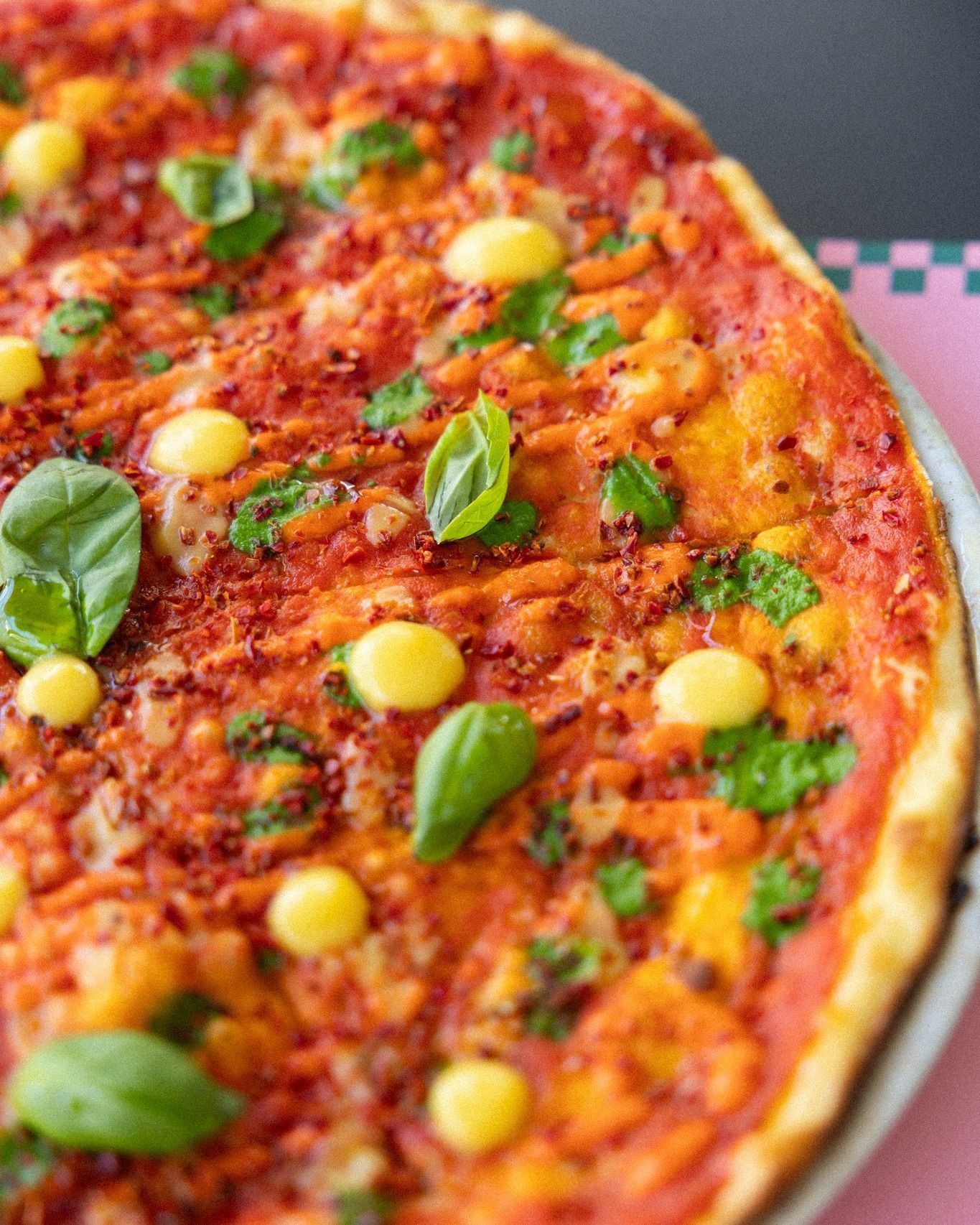 Pizza con salsa di pomodoro, ciuffi gialli e foglie di basilico su una superficie rosa.