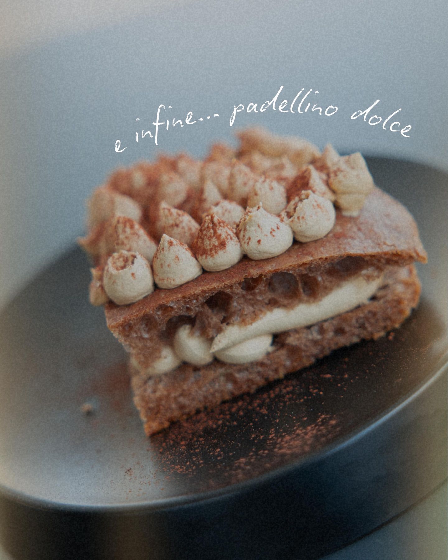 Strati di torta al cioccolato con ripieno di crema, ricoperti di panna montata e cacao in polvere, su un piatto nero.