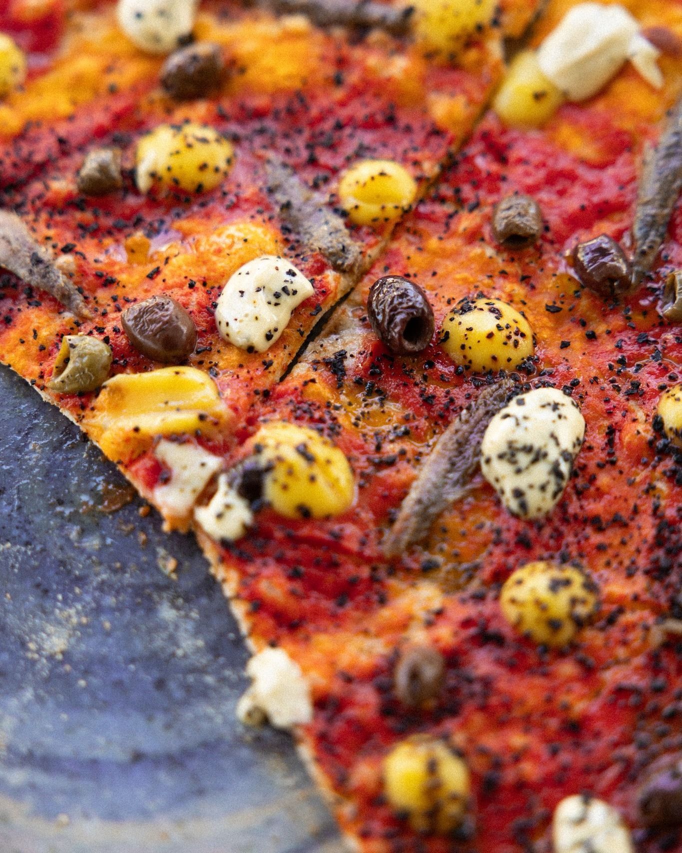 Primo piano di pizza con olive, acciughe, condimenti gialli e bianchi, su un piatto blu.