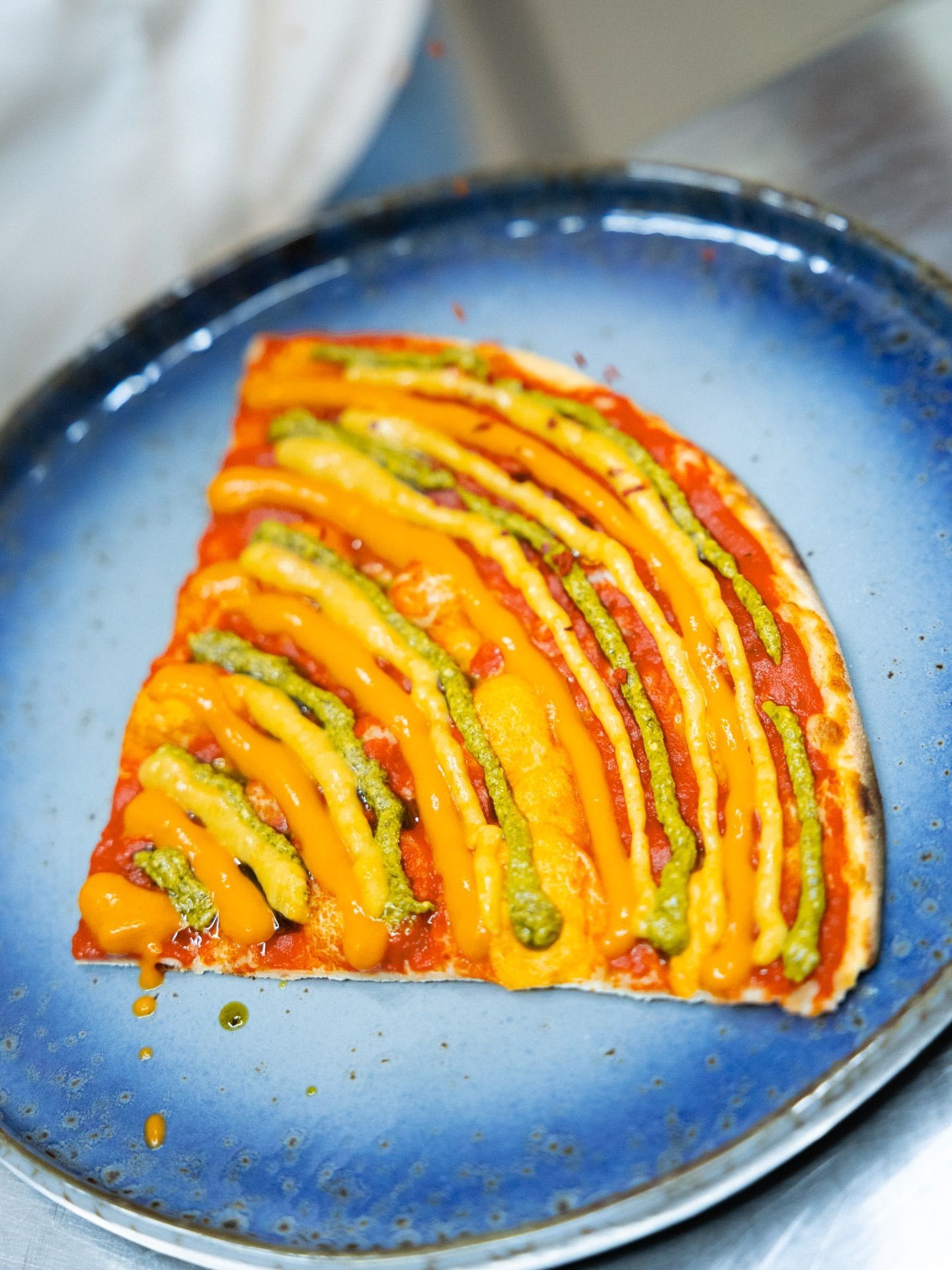 Fetta di pizza su un piatto blu, guarnita con salse arancioni, verdi e gialle in un disegno a strati.
