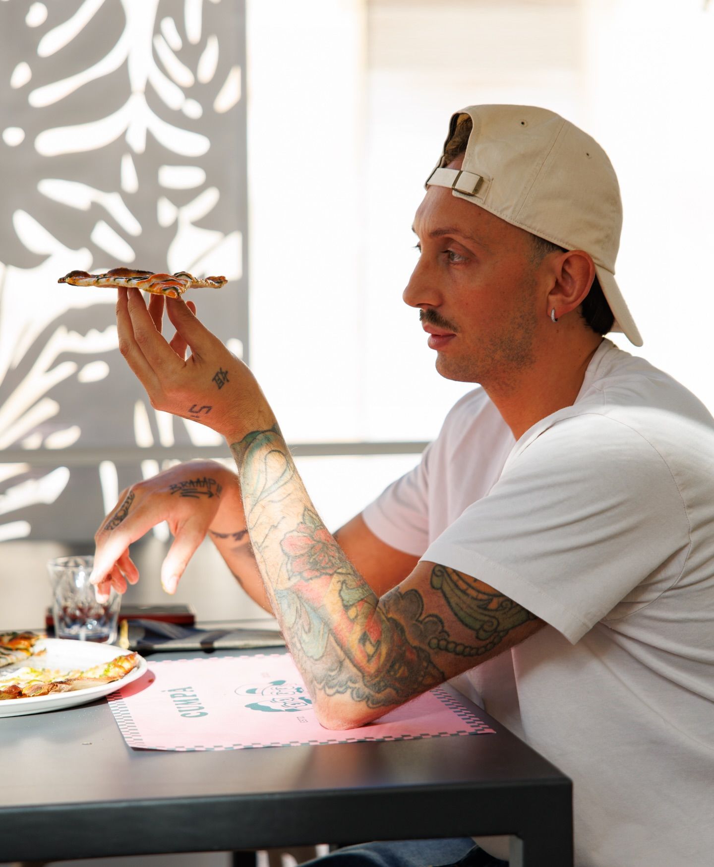 Un uomo con i tatuaggi esamina un pezzo di pizza alla luce del sole, seduto a un tavolo con una bevanda e una tovaglietta rosa.