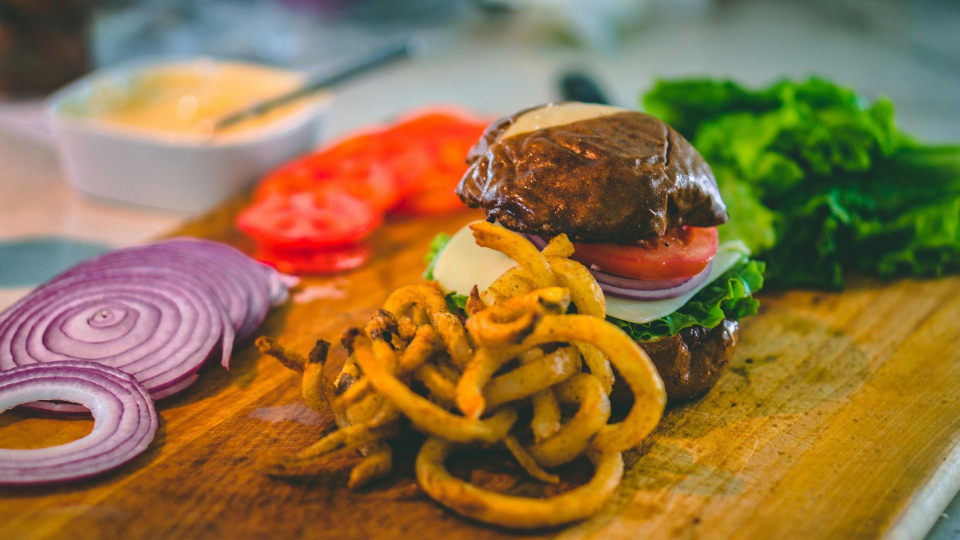 A hamburger with french fries , onions , lettuce and tomatoes on a wooden cutting board.