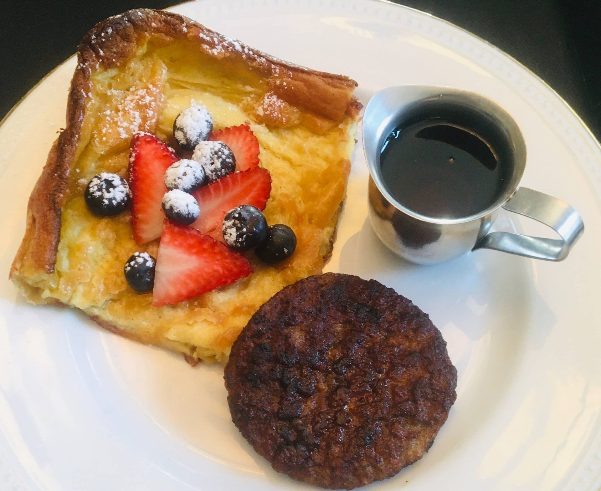 A white plate topped with french toast a sausage and a cup of syrup