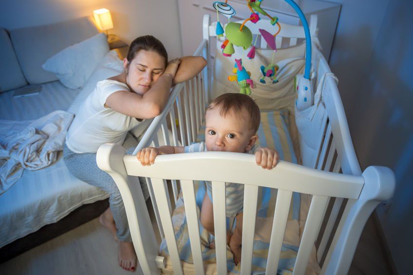Tired mother sleeping on baby's crib while the baby is wide awake