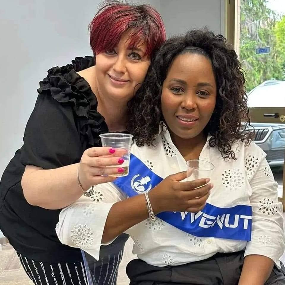 Due donne posano per una foto, una delle quali indossa una fascia con la scritta venut