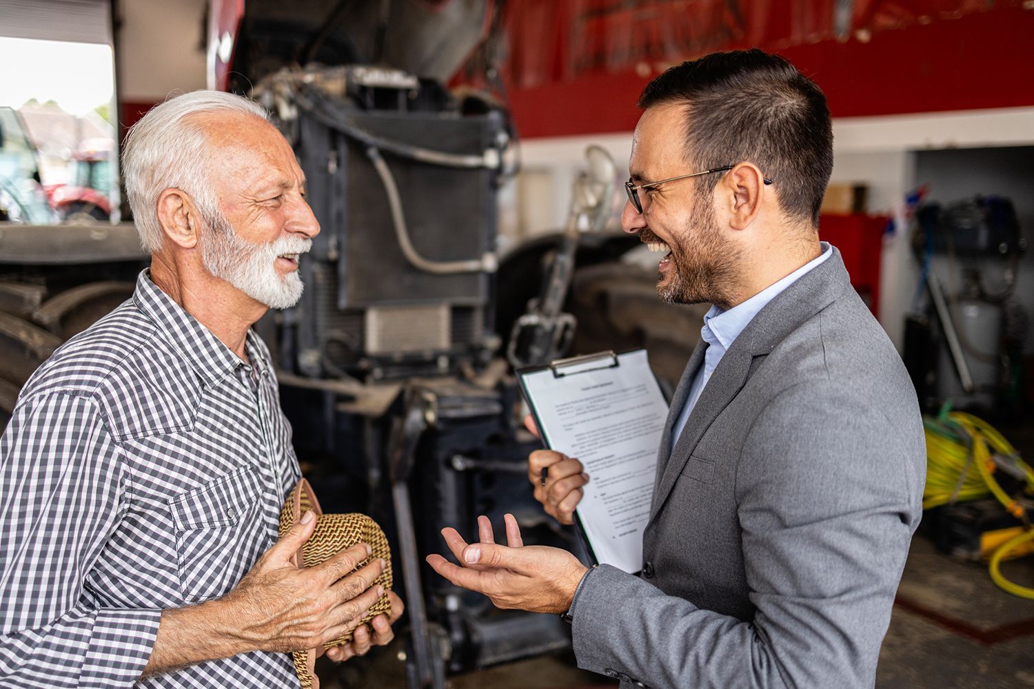 Older man and younger man in front of a tractor, talking. Younger man holds papers, smiling.