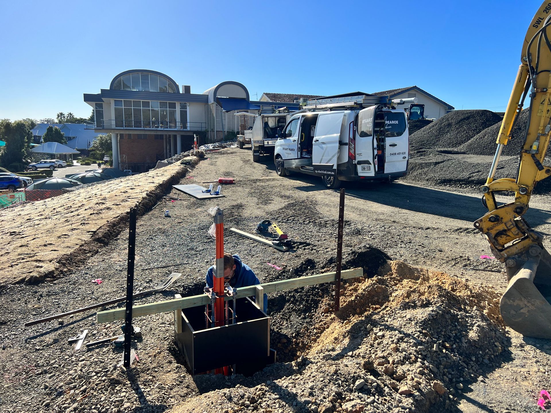 Construction site: worker in a trench with equipment, vans, and a partly-built house under a blue sky — Stuart Pearce Electrical In Coffs Harbour, NSW