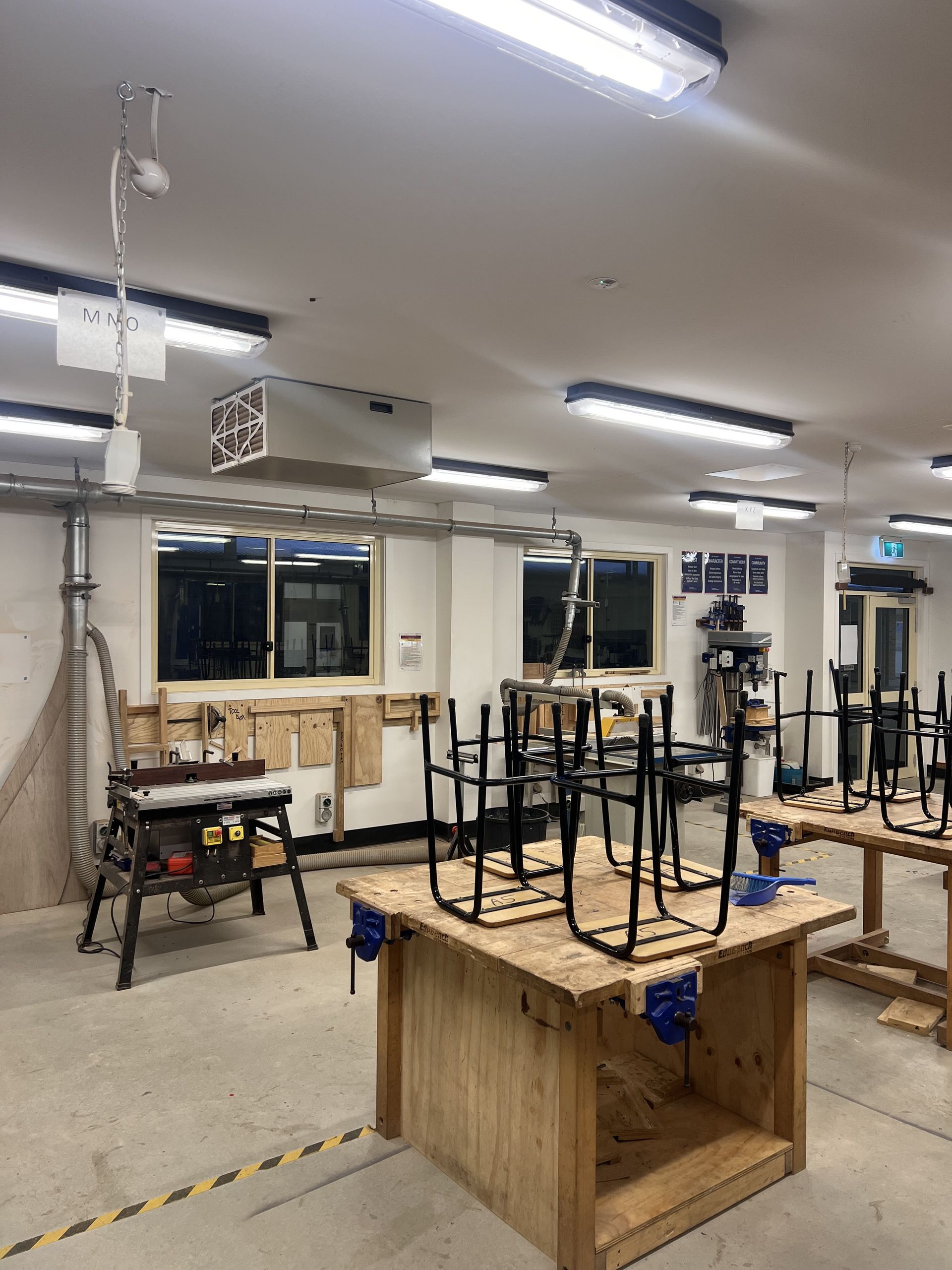 Workshop interior with workbenches, tools, and chairs. Fluorescent lights and a ventilation system are visible — Stuart Pearce Electrical In Coffs Harbour, NSW
