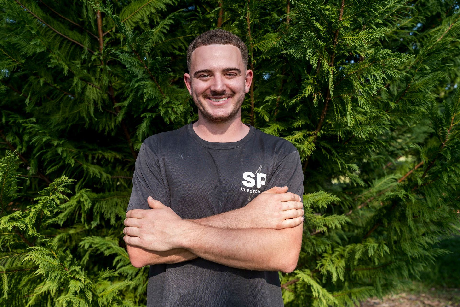 Man with arms crossed smiling in front of green foliage — Stuart Pearce Electrical in Coffs Harbour, NSW