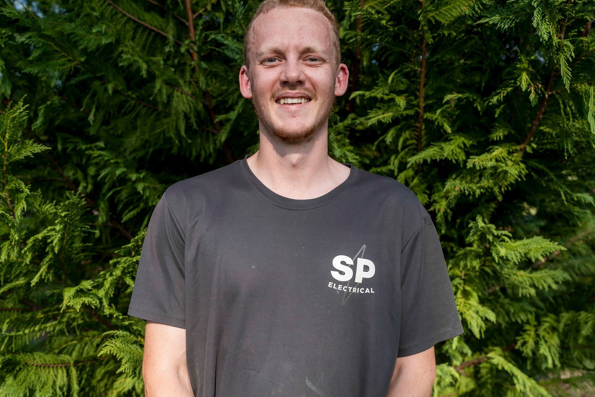 Young man with blonde hair and a black t-shirt smiling in front of green foliage — Stuart Pearce Electrical in Coffs Harbour, NSW