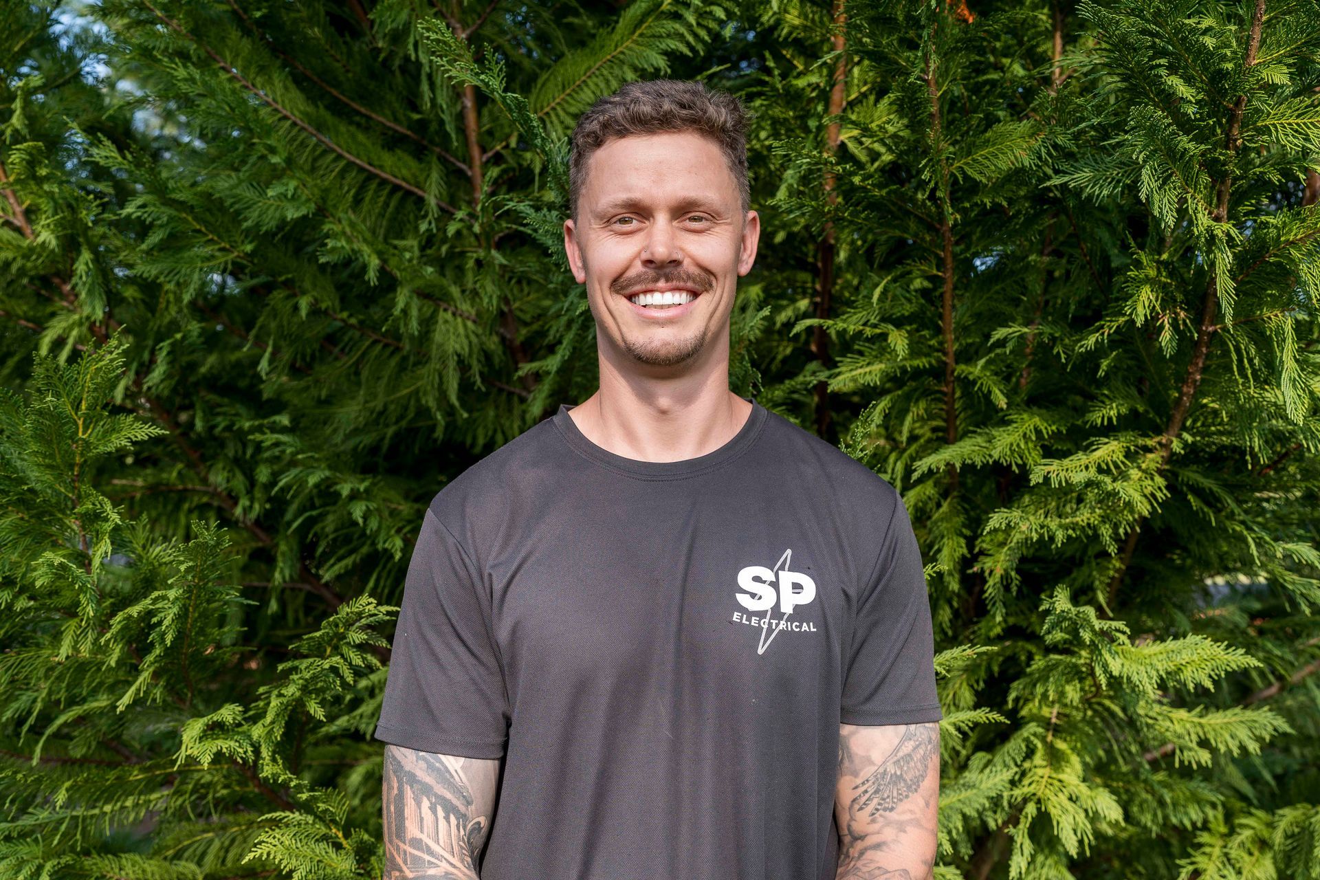 Man smiling, standing in front of green bushes. He wears a black t-shirt with a logo — Stuart Pearce Electrical in Coffs Harbour, NSW
