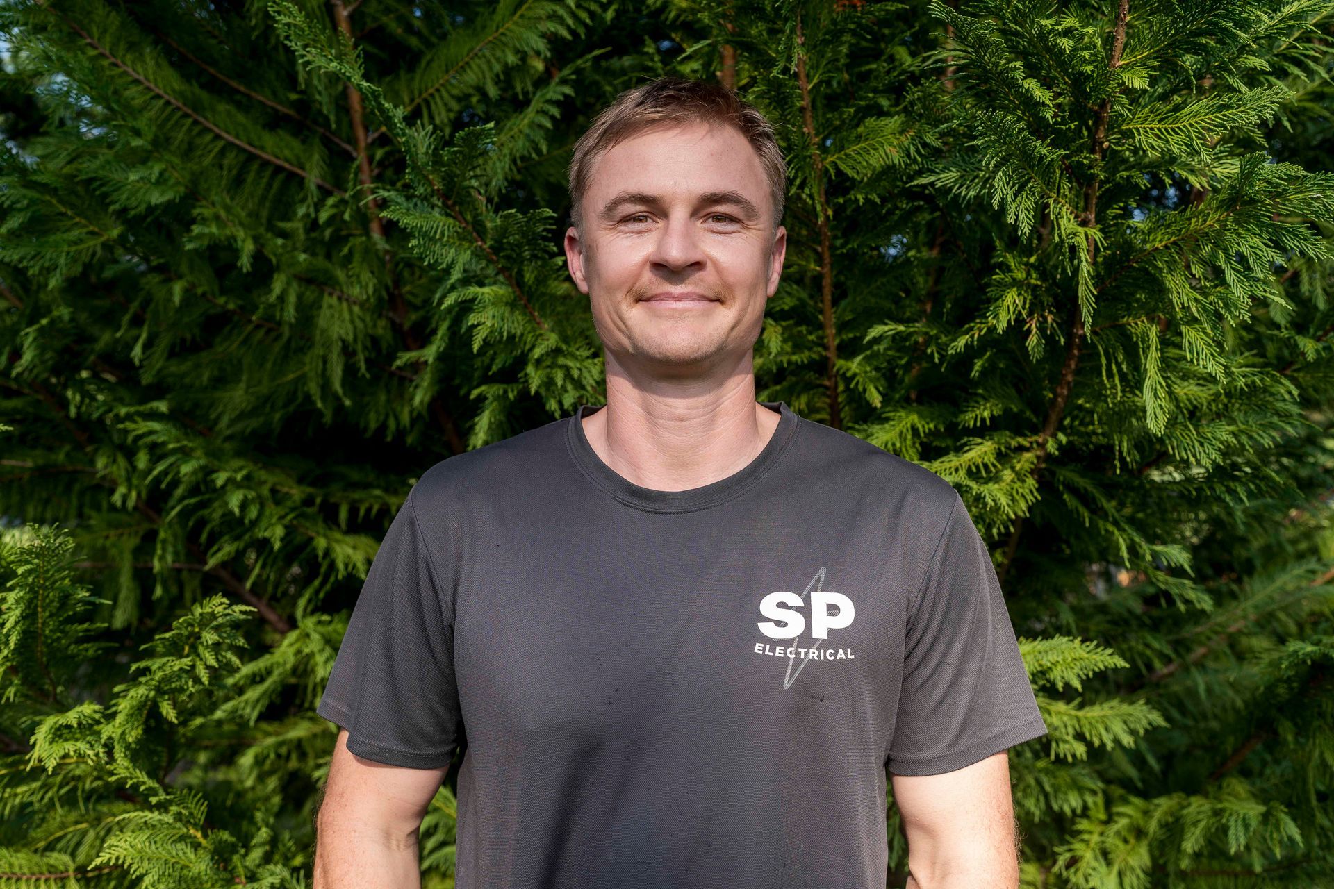 Man in a gray t-shirt with logo smiling in front of a green leafy background — Stuart Pearce Electrical in Coffs Harbour, NSW