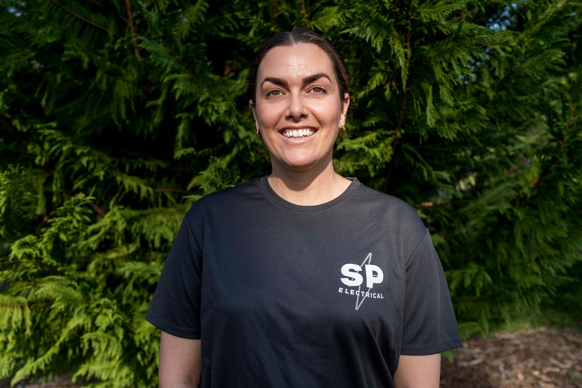 Woman wearing a black shirt with a logo, smiling, in front of green foliage — Stuart Pearce Electrical in Coffs Harbour, NSW