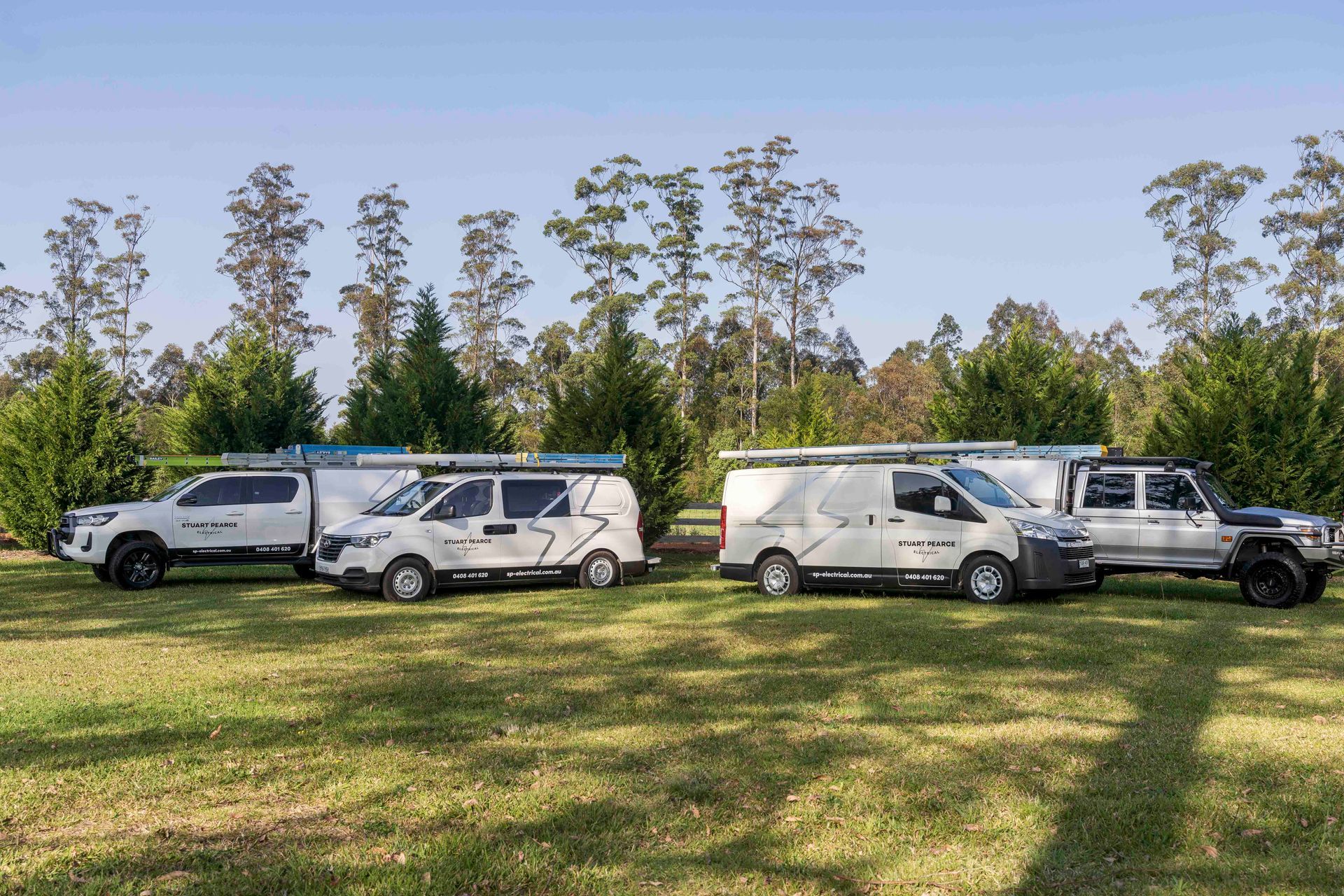 Four white vehicles parked on a grassy field with trees — Stuart Pearce Electrical In Coffs Harbour, NSW