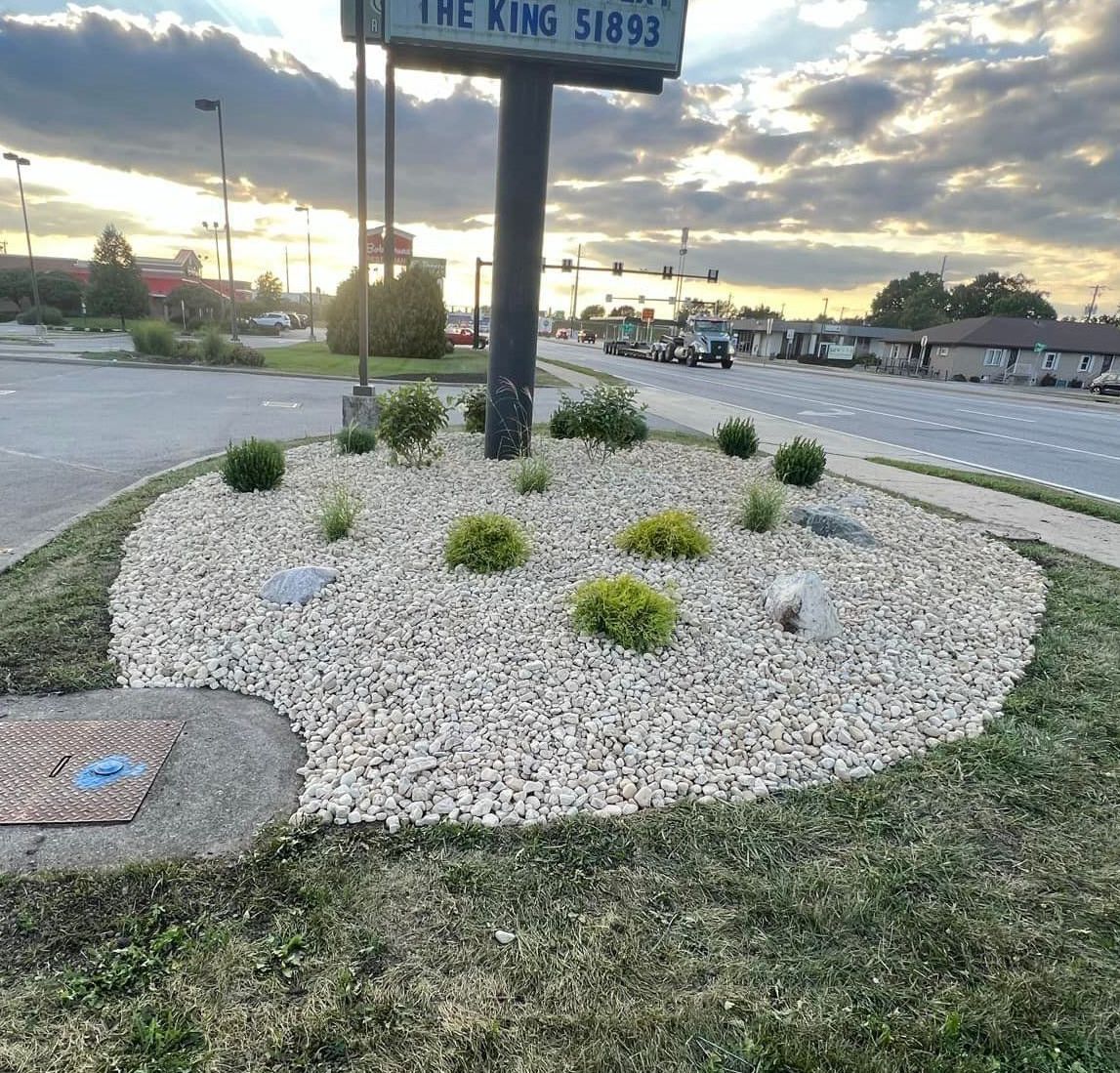 A gravel-covered flower bed with green shrubs surrounds a signpost. The setting is a parking lot or median with a street and buildings in the background.