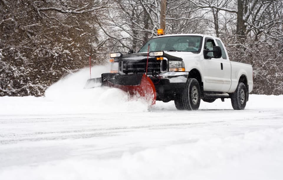 White snow plow truck clearing a snow-covered road in a wintery landscape.