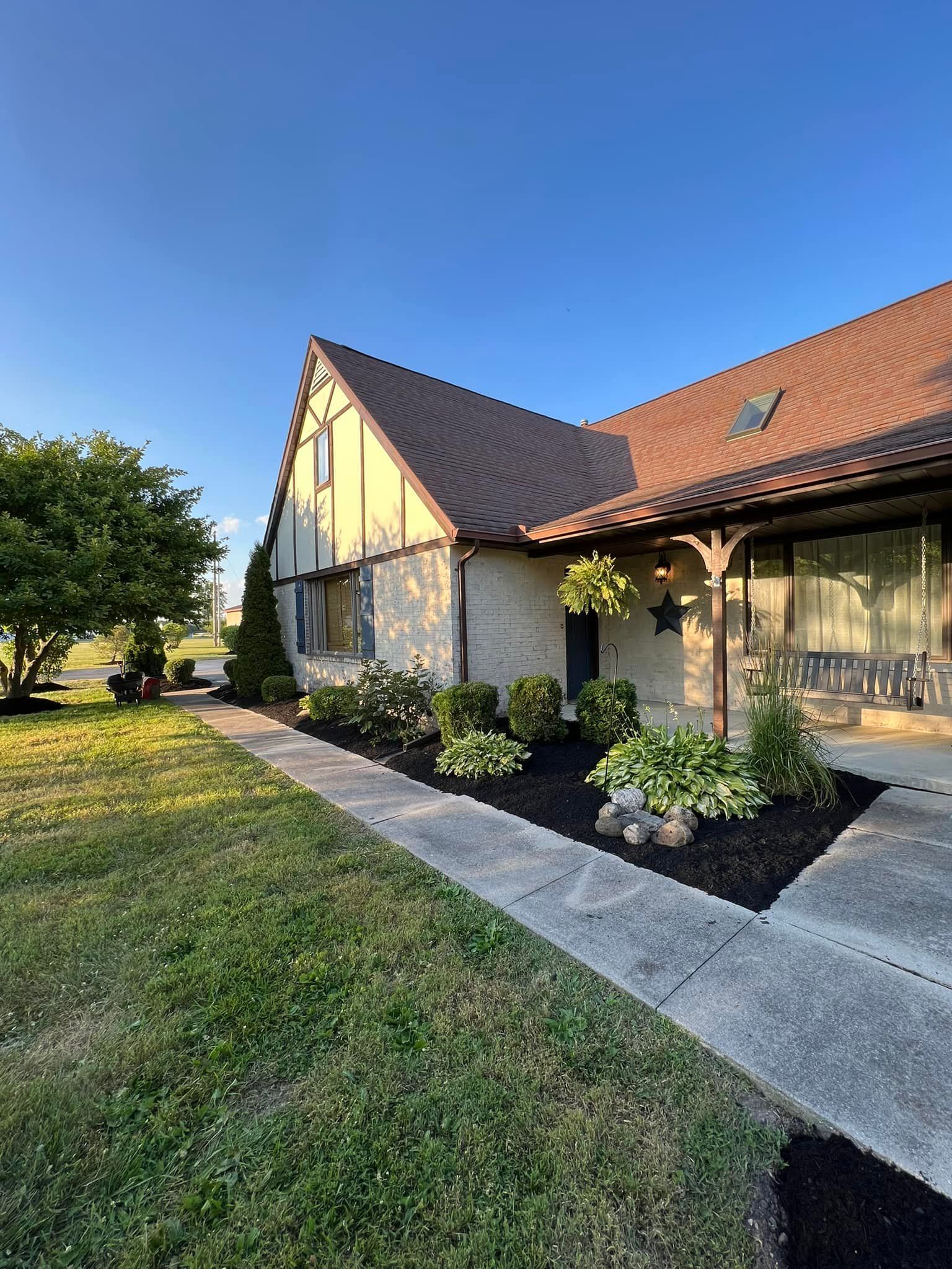 Tudor-style house with tan stucco, brown tile roof, and landscaped front yard. Blue sky background.