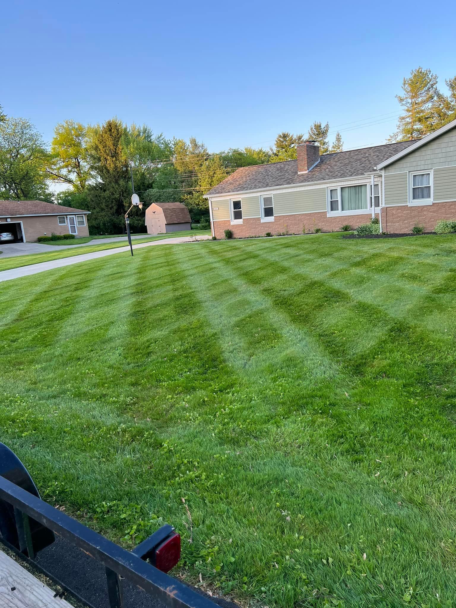 A neatly mowed lawn in a suburban setting with a house, trees, and a basketball hoop.